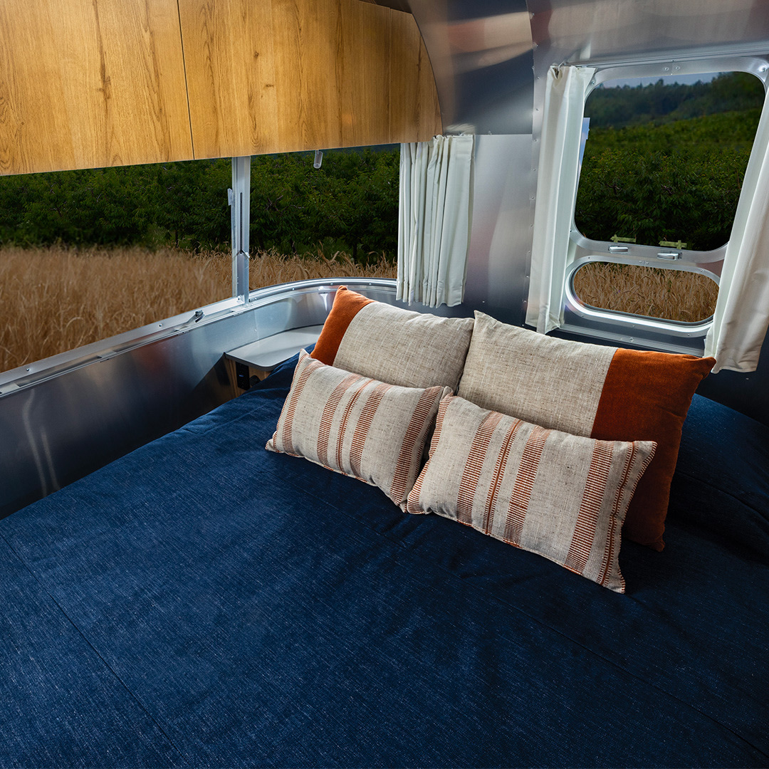 Cozy RV rental bed with pillows, offering scenic views of golden fields and green trees outside. Perfect for camping adventures.. Airstream Trade Wind 2025