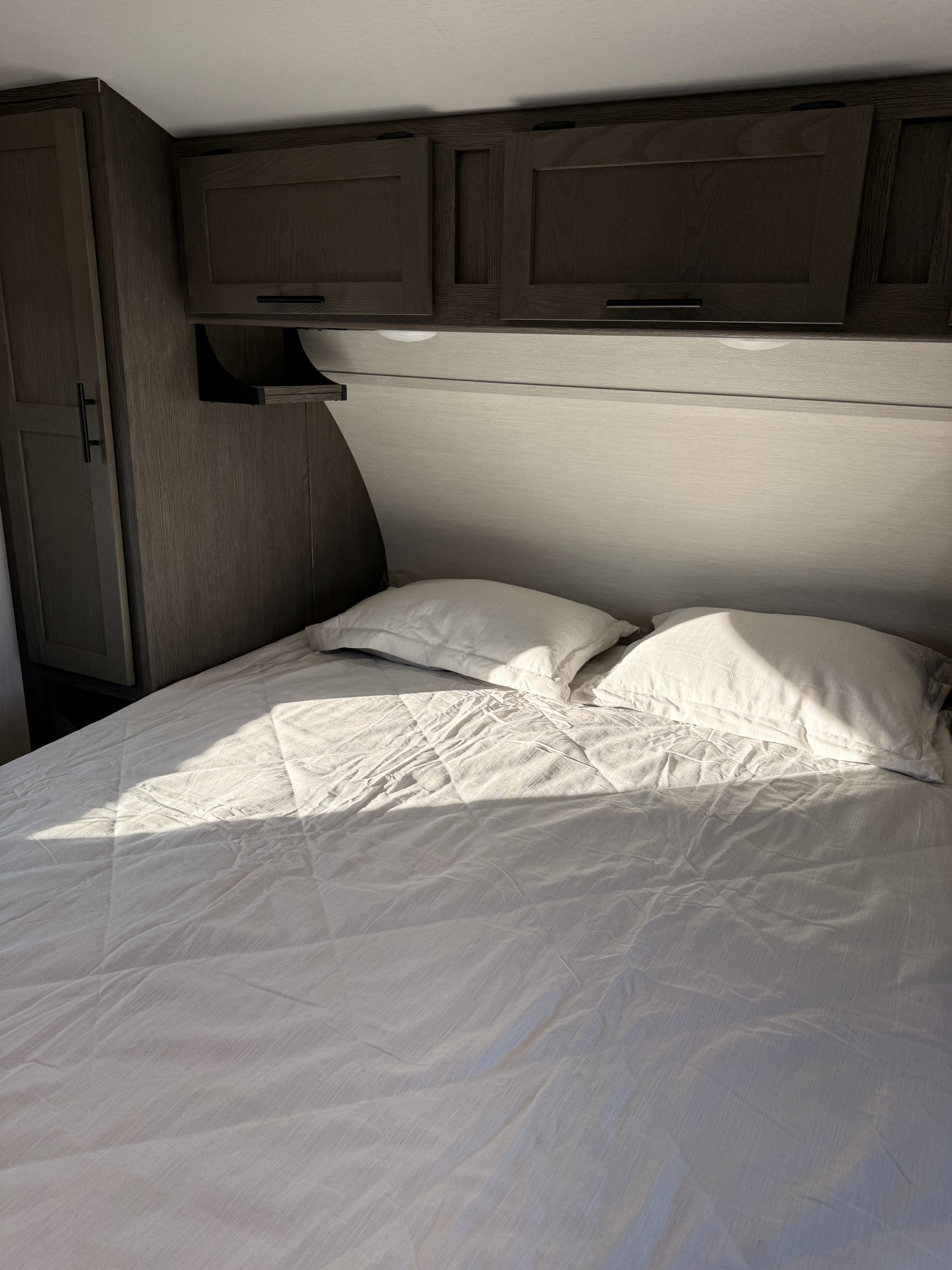 Cozy RV rental bedroom with white bedding, pillows, and dark wood cabinets above. Sunlight streams across the bed.. Forest River Artic Wolf 2850PT 2026