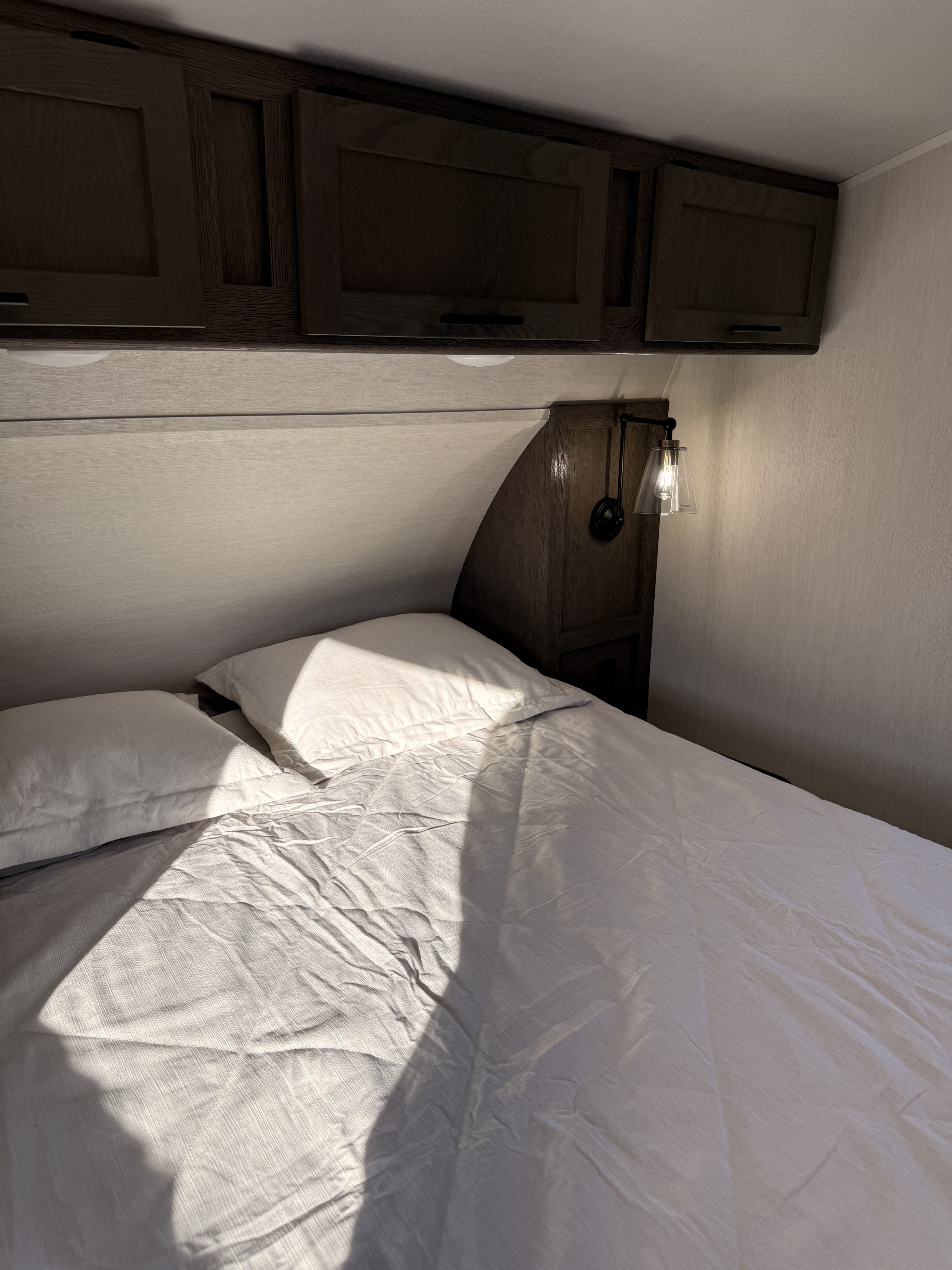 A comfortable RV rental bedroom featuring a bed with white pillows and duvet, overhead storage cabinets, and a bedside lamp.. Forest River Artic Wolf 2850PT 2026