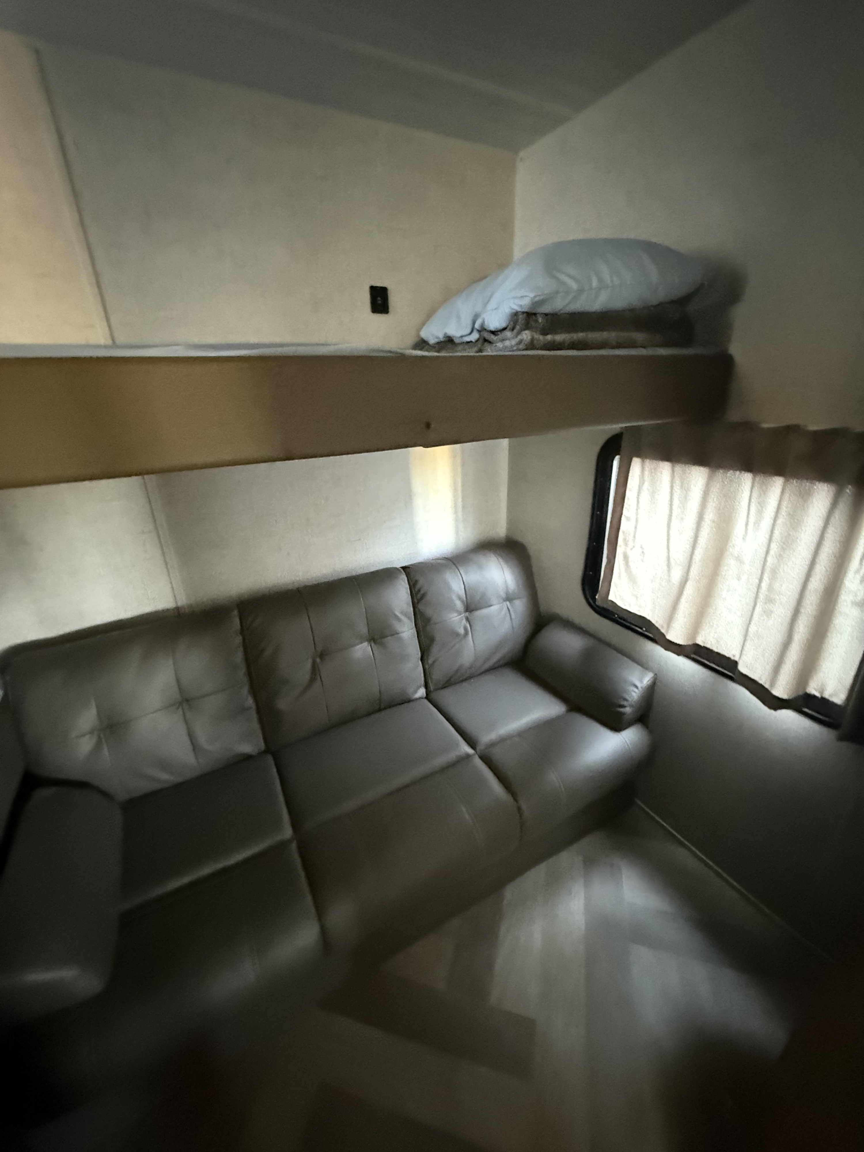 Comfortable RV rental interior: a brown sofa, an upper bunk bed with a pillow, and a curtained window create a cozy space.. Forest River Wildwood X-Lite 2021