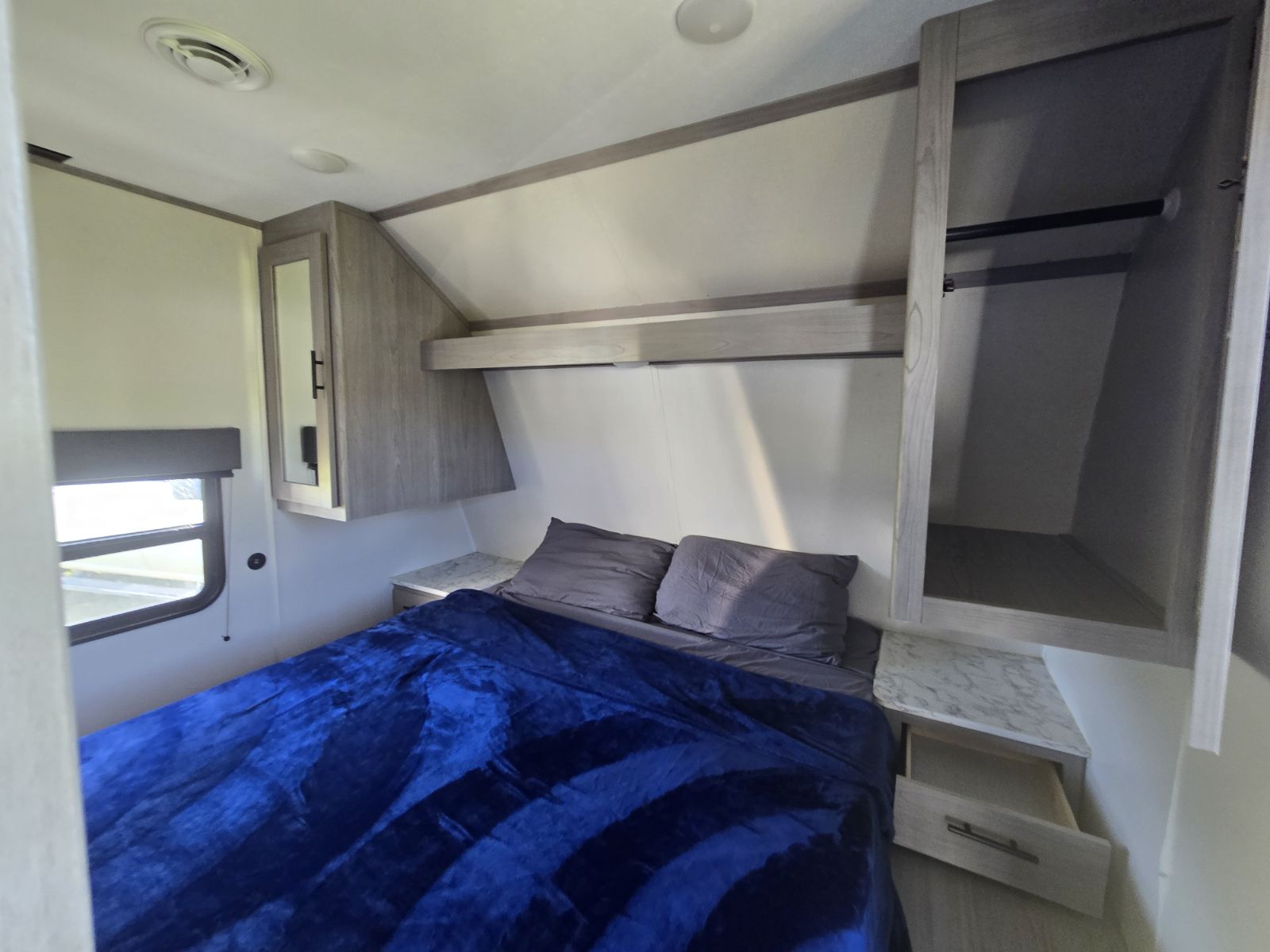 Cozy RV rental bedroom featuring a bed with blue blanket, window, ceiling vents, and wood storage cabinets with a closet.. Coleman Lantern LT 26BH 2022