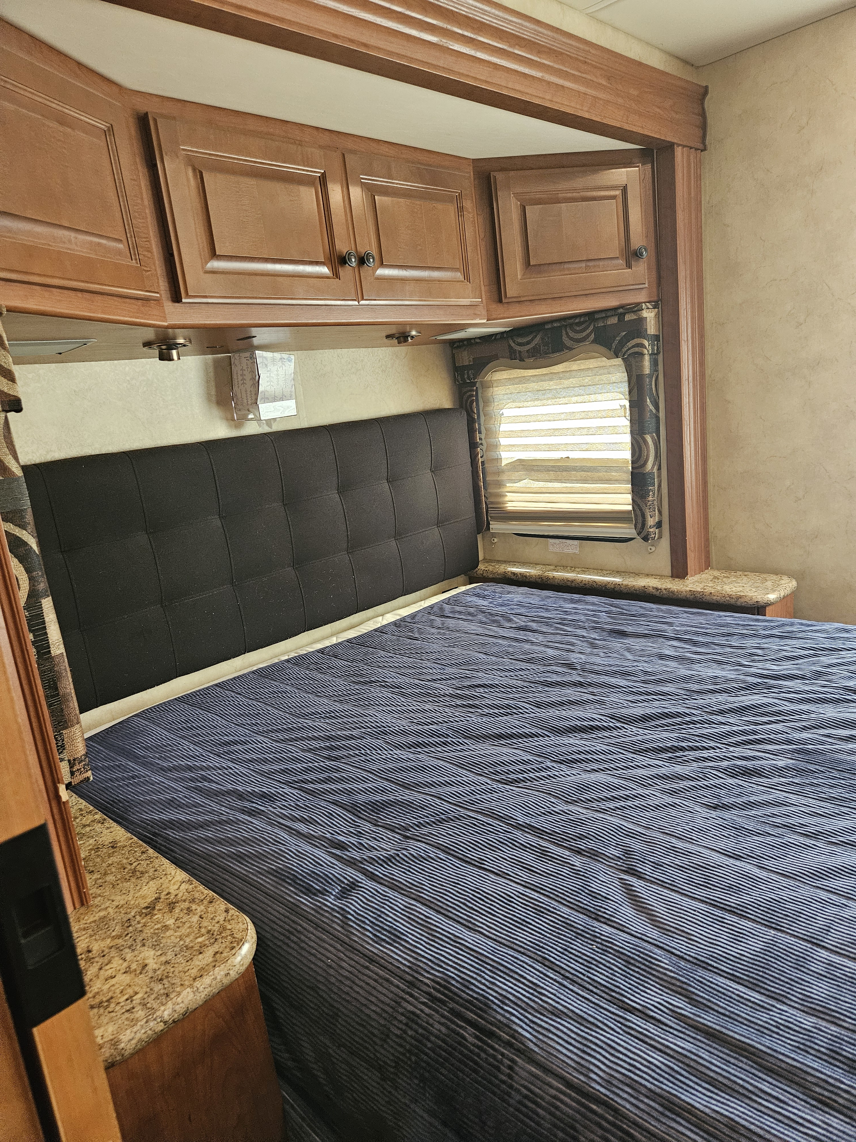 This RV rental bedroom offers a cozy bed with a dark blue quilted spread, black headboard, wooden cabinets, and a window with blinds.. Four Winds Windsport 2011