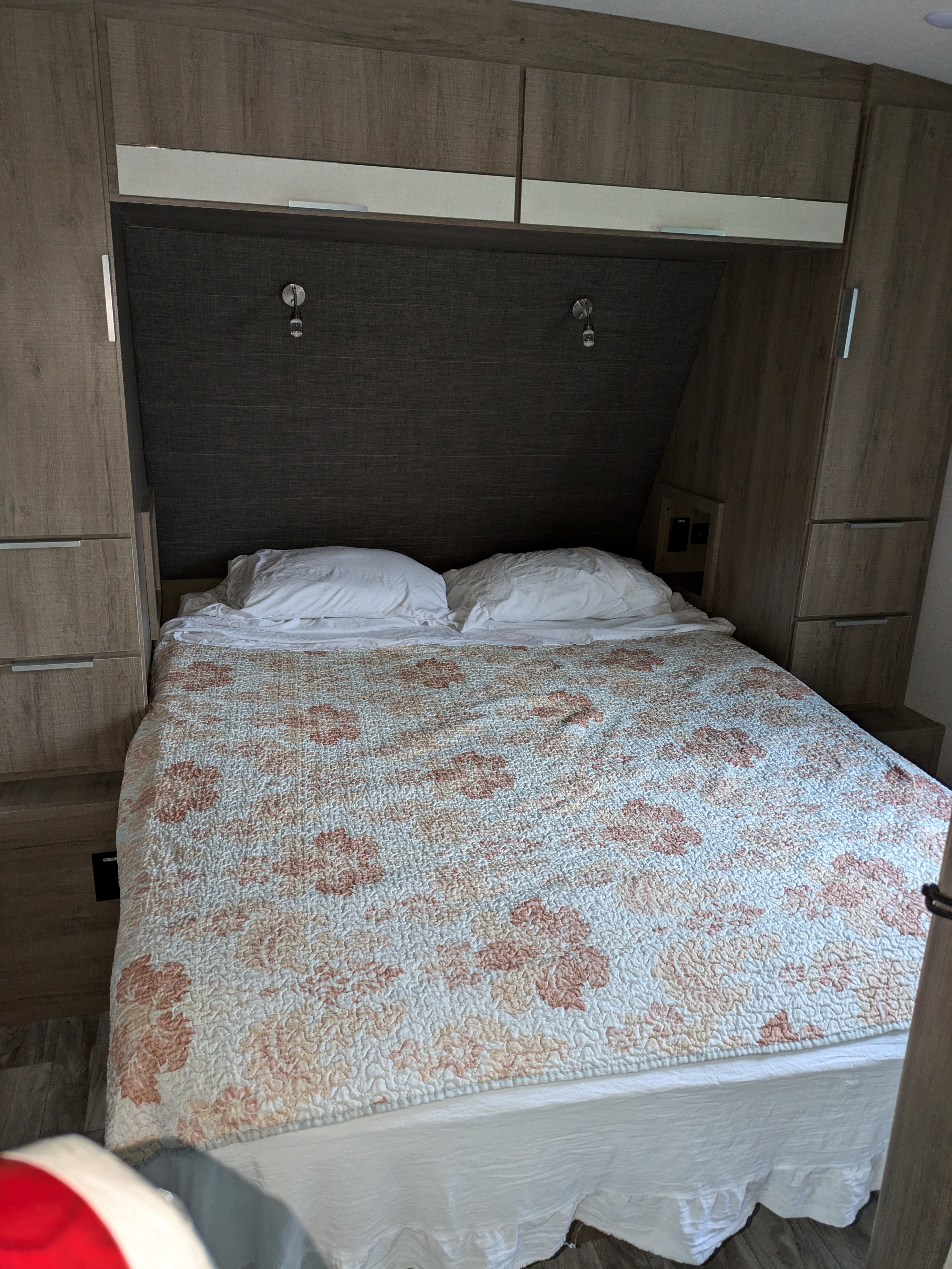Inside an RV rental: a compact bedroom with a bed featuring a floral quilt, white pillows, and wood cabinets.. Grand Design Imagine 2021