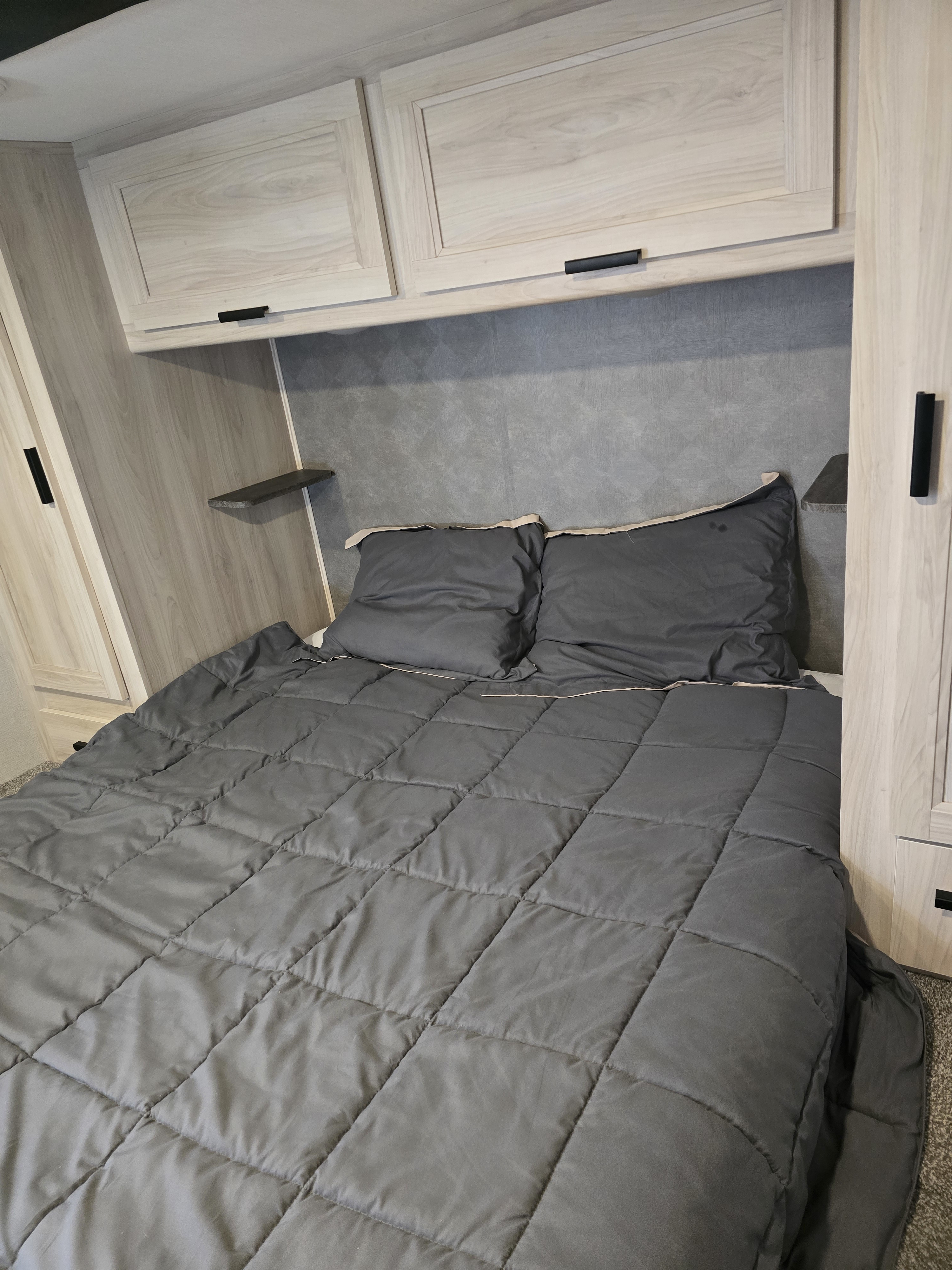 Inside a modern rv rental: a cozy bedroom with a quilted gray comforter, pillows, light wood cabinets, and shelving.. Forester LE 2023