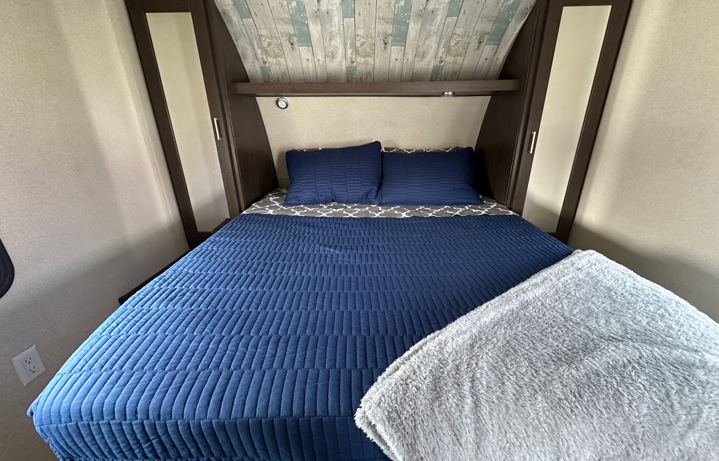 Cozy RV rental bedroom with blue quilted bedding, navy pillows, and a fluffy grey blanket. Features wood trim and plank ceiling.. Forest River Salem 2018