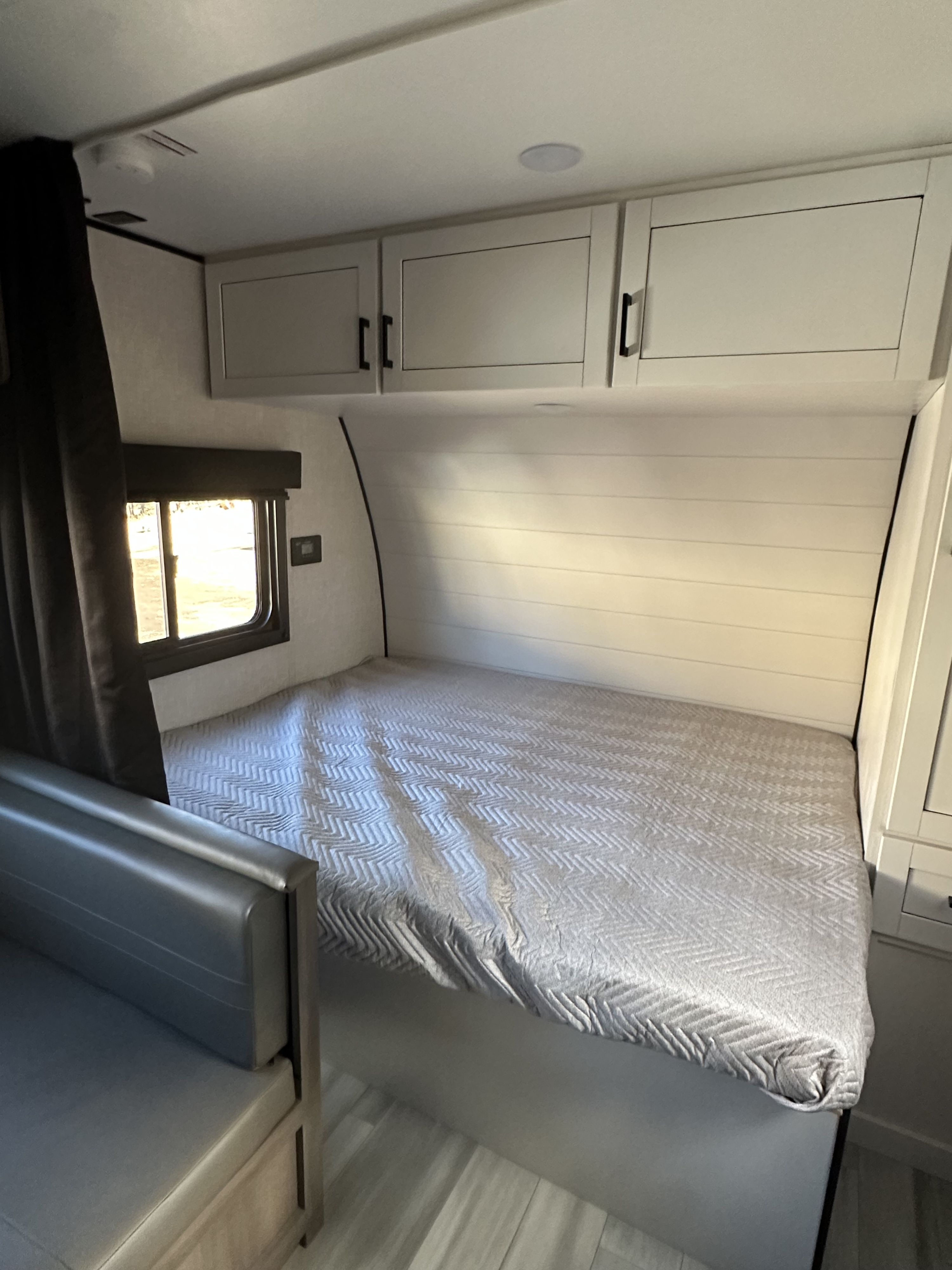 This RV rental features a compact bedroom: a bed with chevron quilt, overhead storage, and a bright window.. Starcraft Autumn Ridge 2025