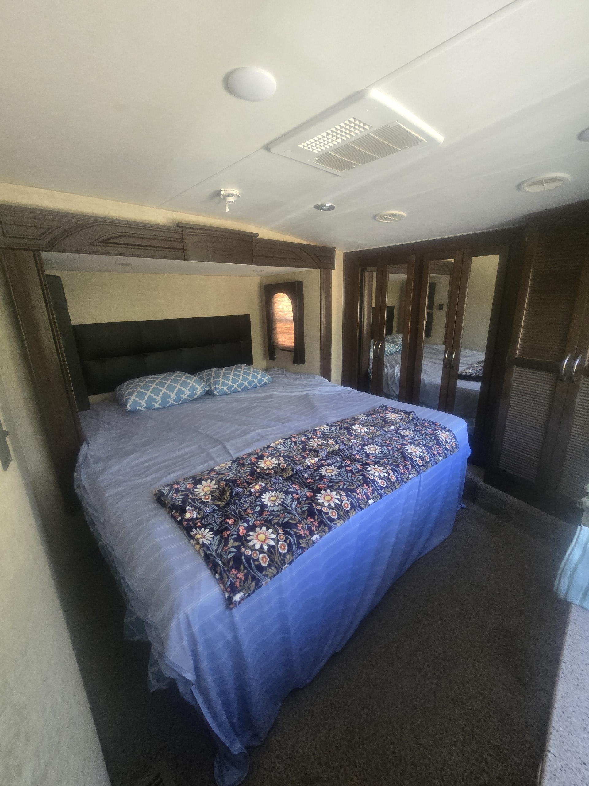 Cozy RV rental bedroom featuring a queen bed with blue bedding, mirrored closets, and a small window. Ideal RV accommodation.. Heartland RVs Cyclone HD Edition Toy Hauler 2015