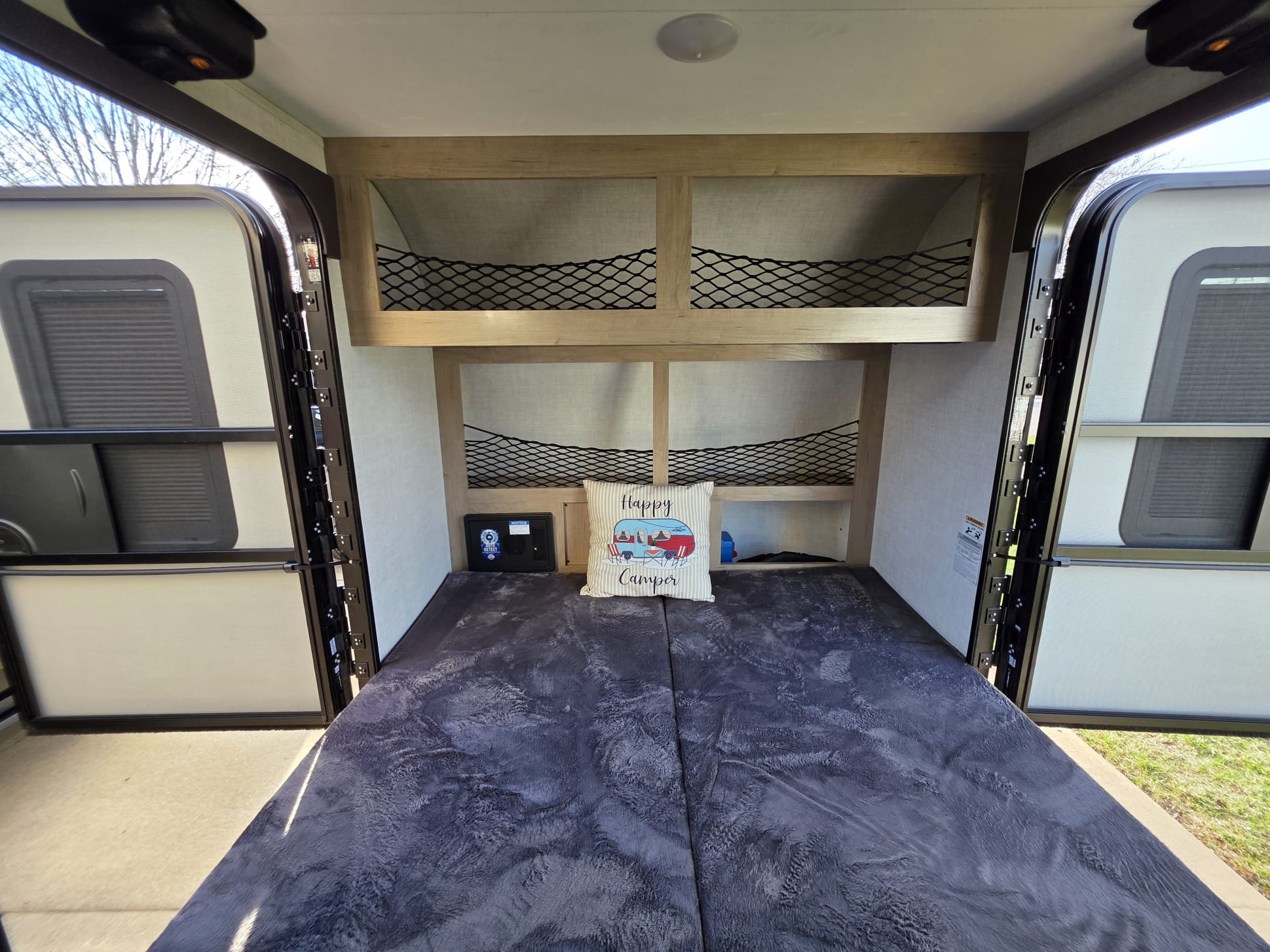 This RV rental shows a compact interior with a bed, "Happy Camper" pillow, overhead storage with netting, and open back doors.. Ember E Series Spark LE 2026