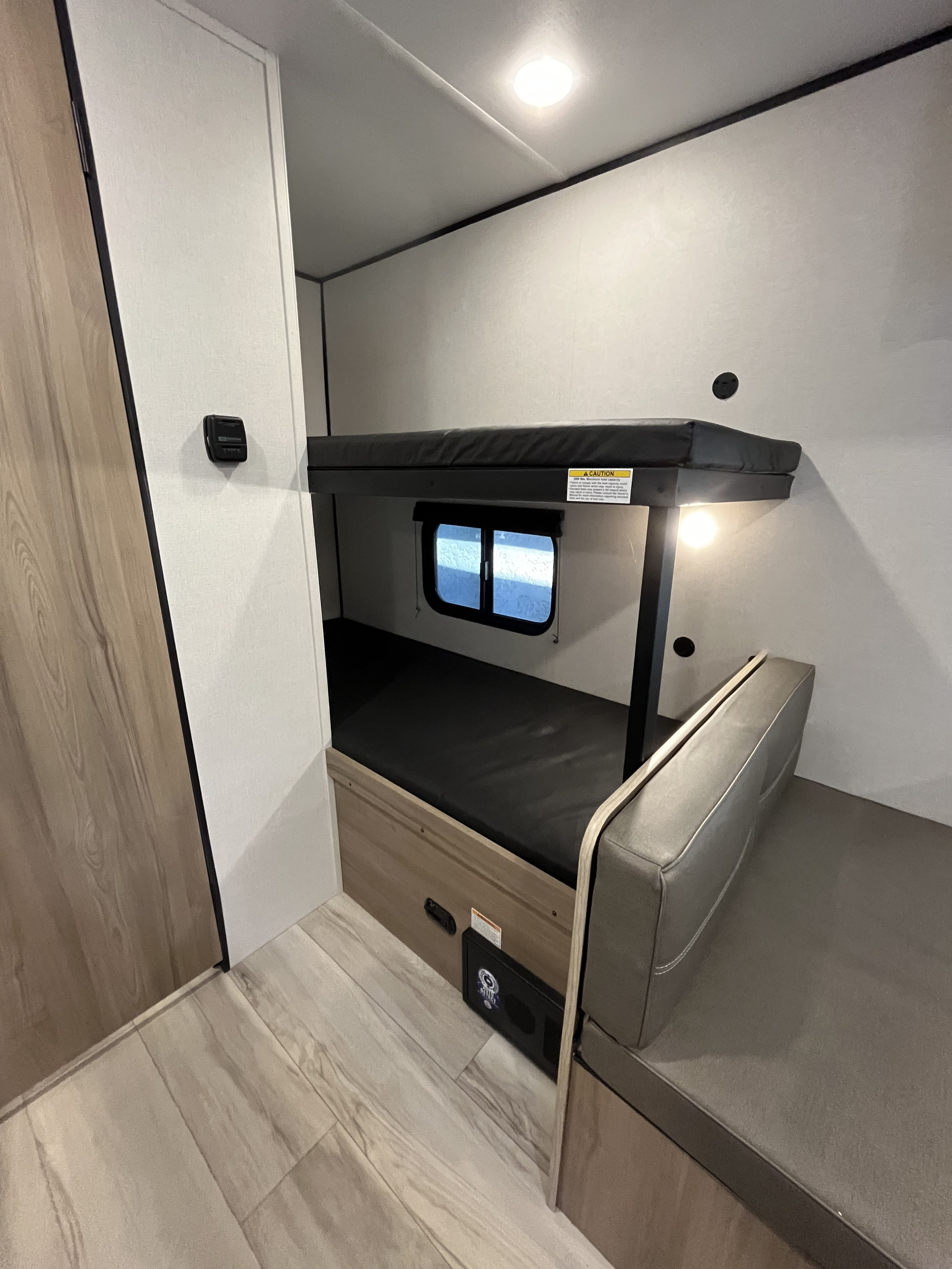 Cozy RV rental bunk bed area: light walls, dark mattresses, window, bright light, and grey dinette bench.. Keystone RV Coleman 2025