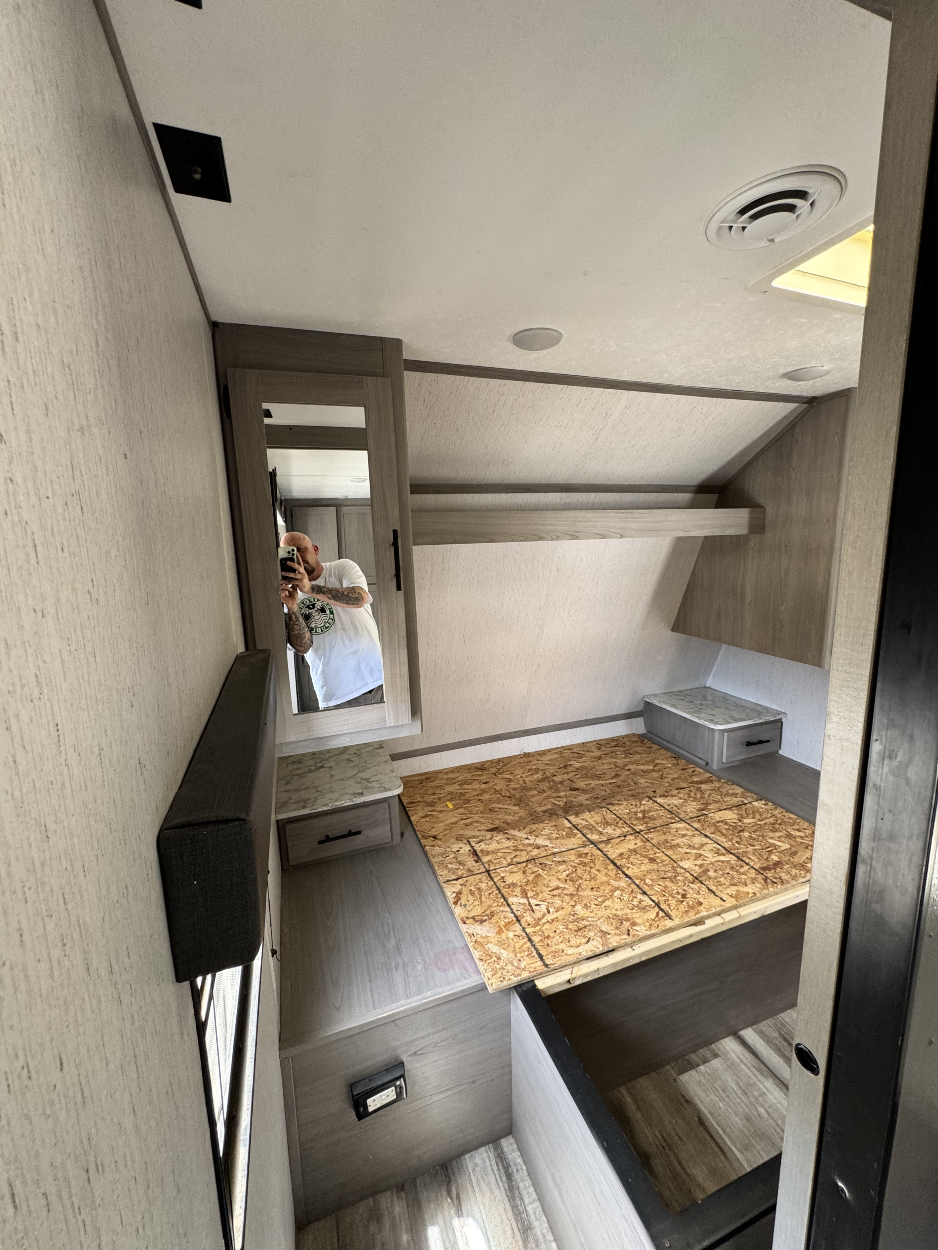 RV rental bedroom: OSB bed platform, nightstands, and overhead storage are visible. A person is reflected in the mirror.. Coleman Cch 2022