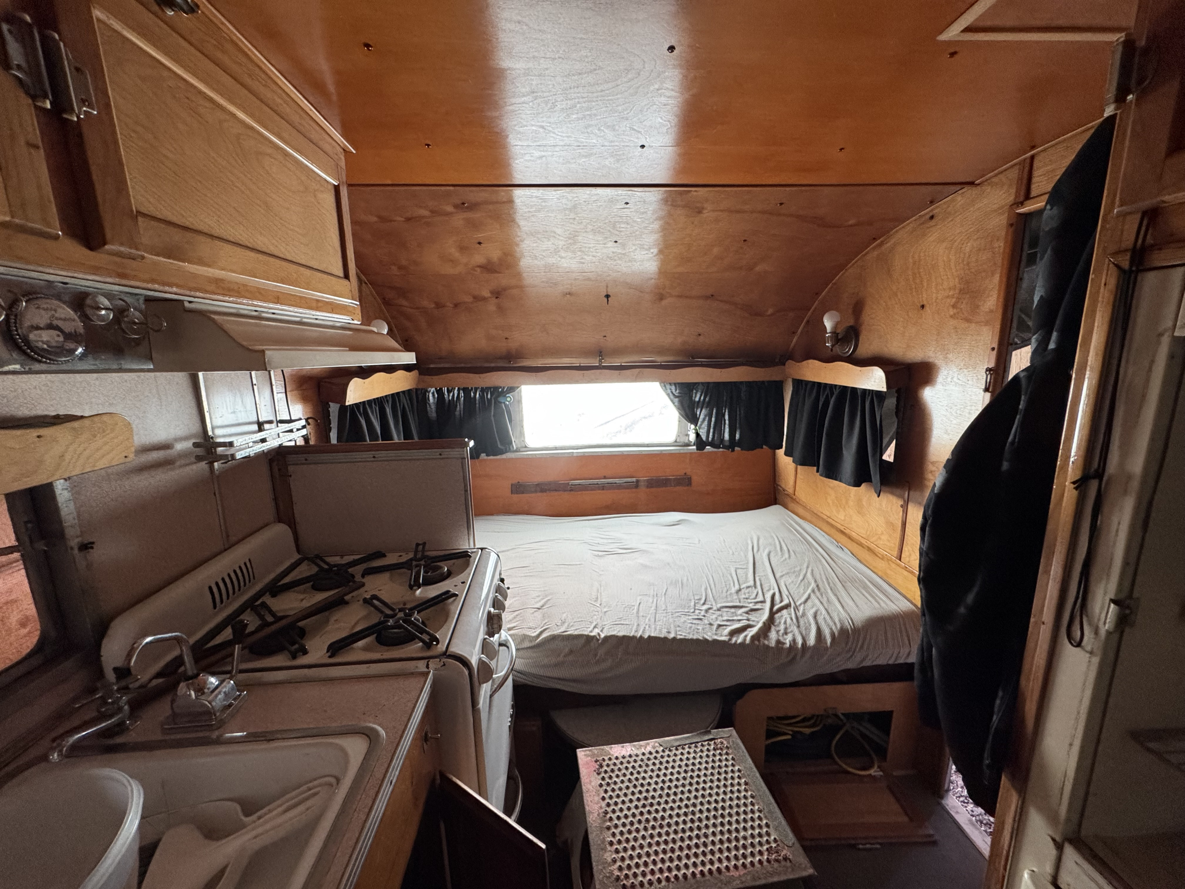 Cozy vintage RV rental interior featuring warm wood paneling, a compact kitchen with stove and sink, and a comfortable bed.. Boles Aero Travel Trailer 1950