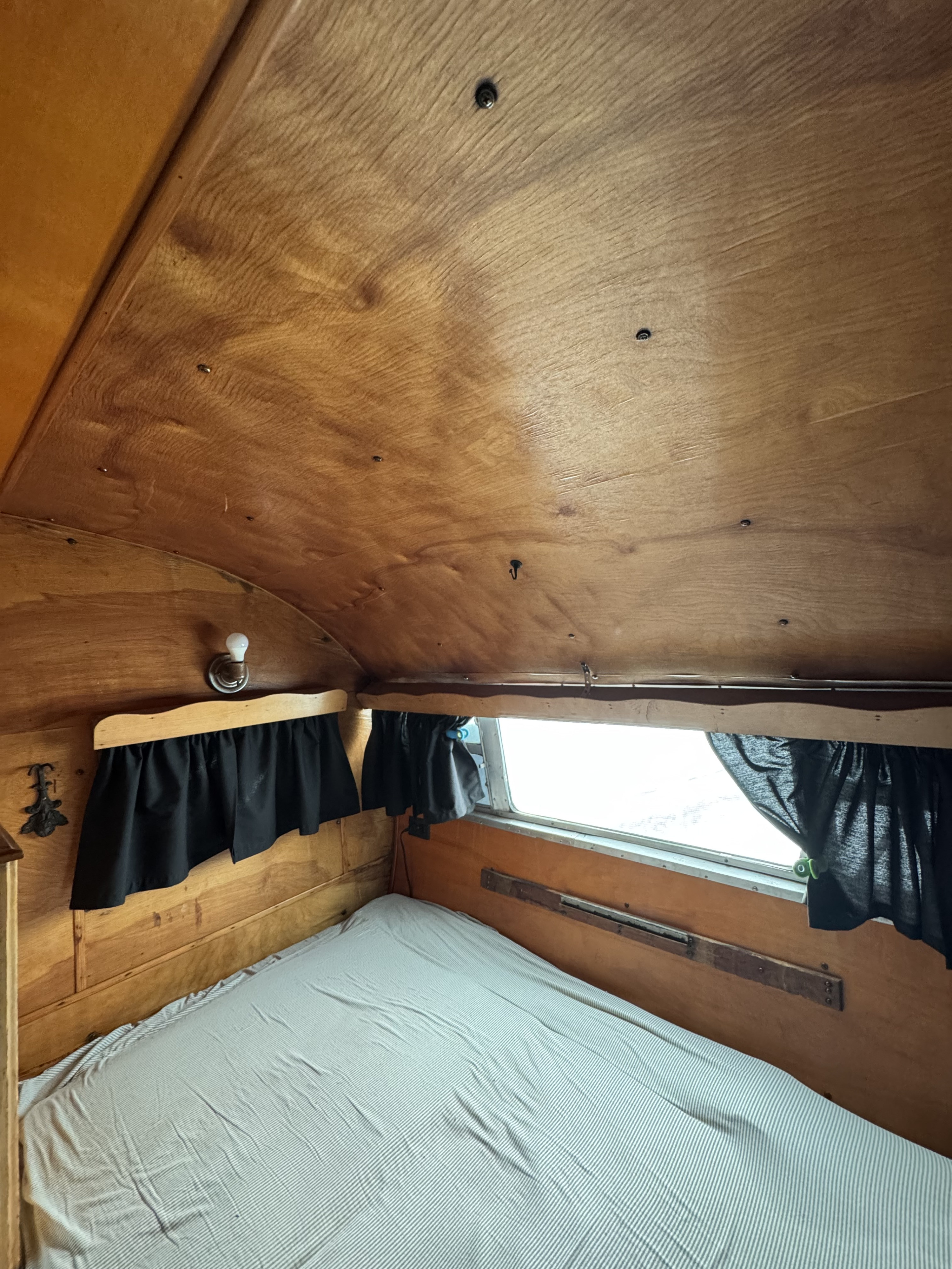 RV rental bedroom: warm wood paneled walls & ceiling, a striped bed, and dark privacy curtains.. Boles Aero Travel Trailer 1950