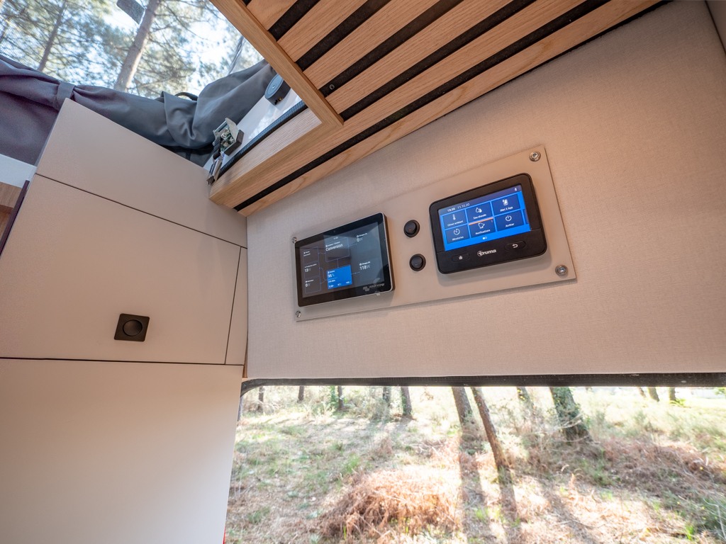 RV rental interior: two digital control panels manage systems, with modern cabinetry & forest views from a large window.. Ford Transit 2024