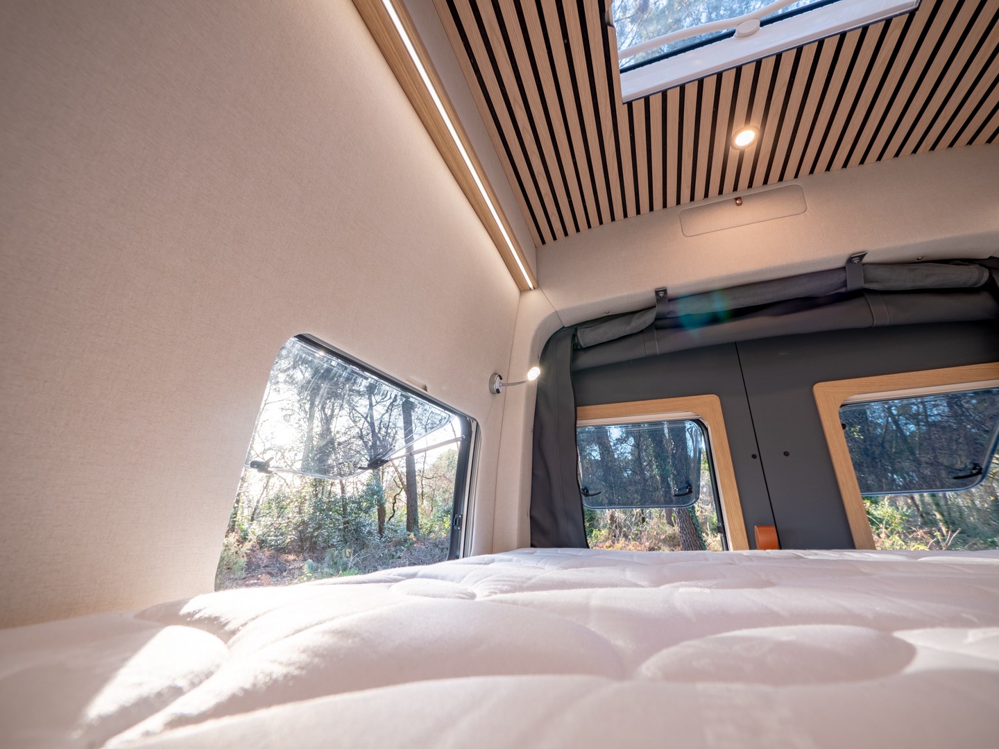 Cozy bed & nature views from a modern RV rental's sleeping area, featuring a skylight, wood-slat ceiling, and ambient lighting.. MAN TGE 4x4 2025