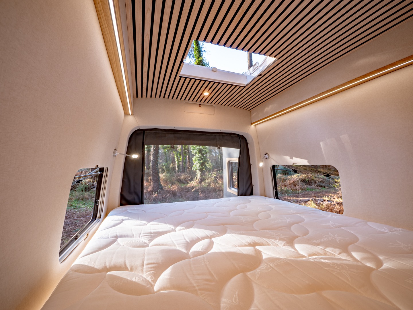 Bright RV rental bedroom interior with a comfy bed, skylight, and windows offering serene forest views. Modern design elements.. MAN TGE 4x4 2025
