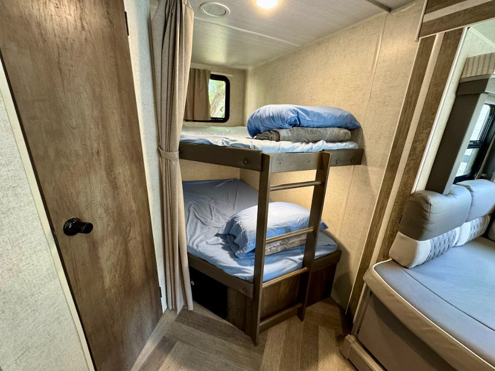 Cozy RV rental bunk beds with blue bedding & ladder, offering compact sleeping space, visible from a door and seating area.. Wildwood 263BHXL 2021