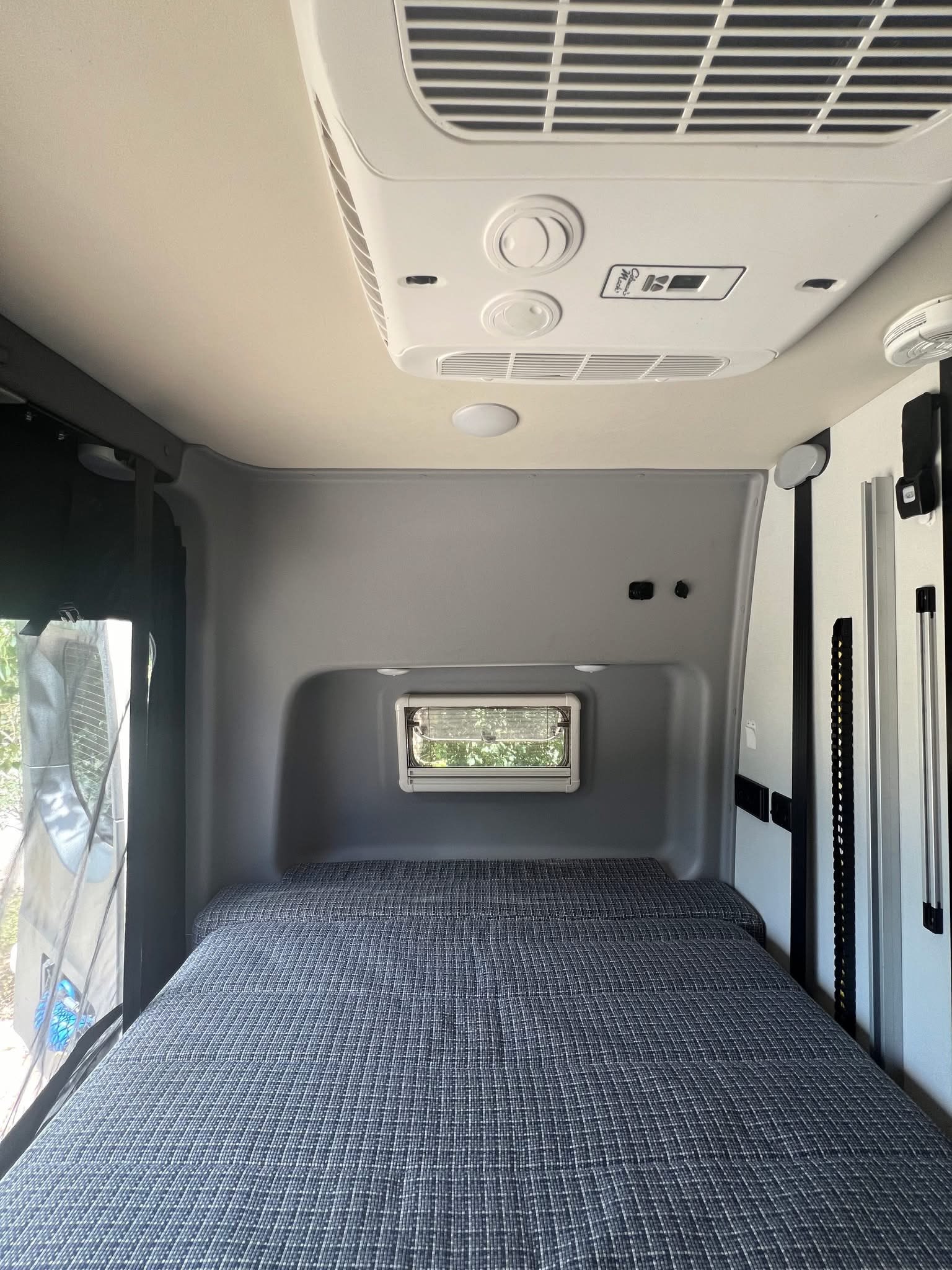 Inside an rv rental, a cozy campervan bedroom features a blue patterned bed, AC unit, and small window for views.. Winnebago Revel 2024