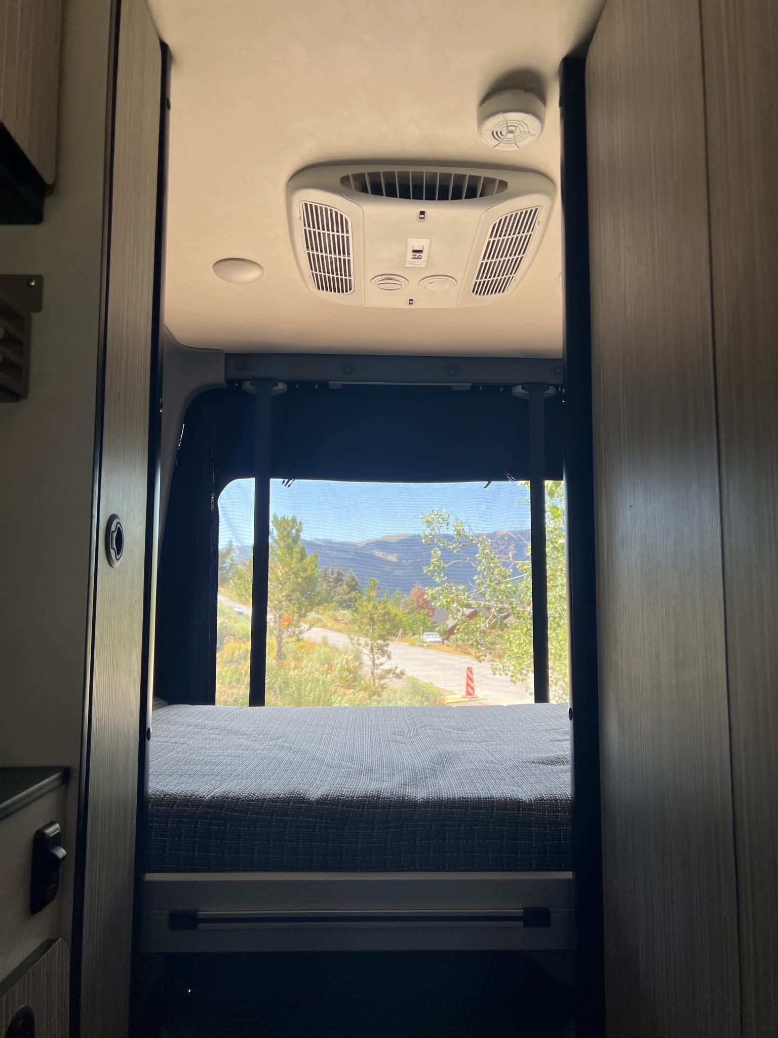 Inside an RV rental, a cozy bed faces a scenic mountain view from a large rear window. Overhead AC and smoke detector.. Winnebago Revel 2024