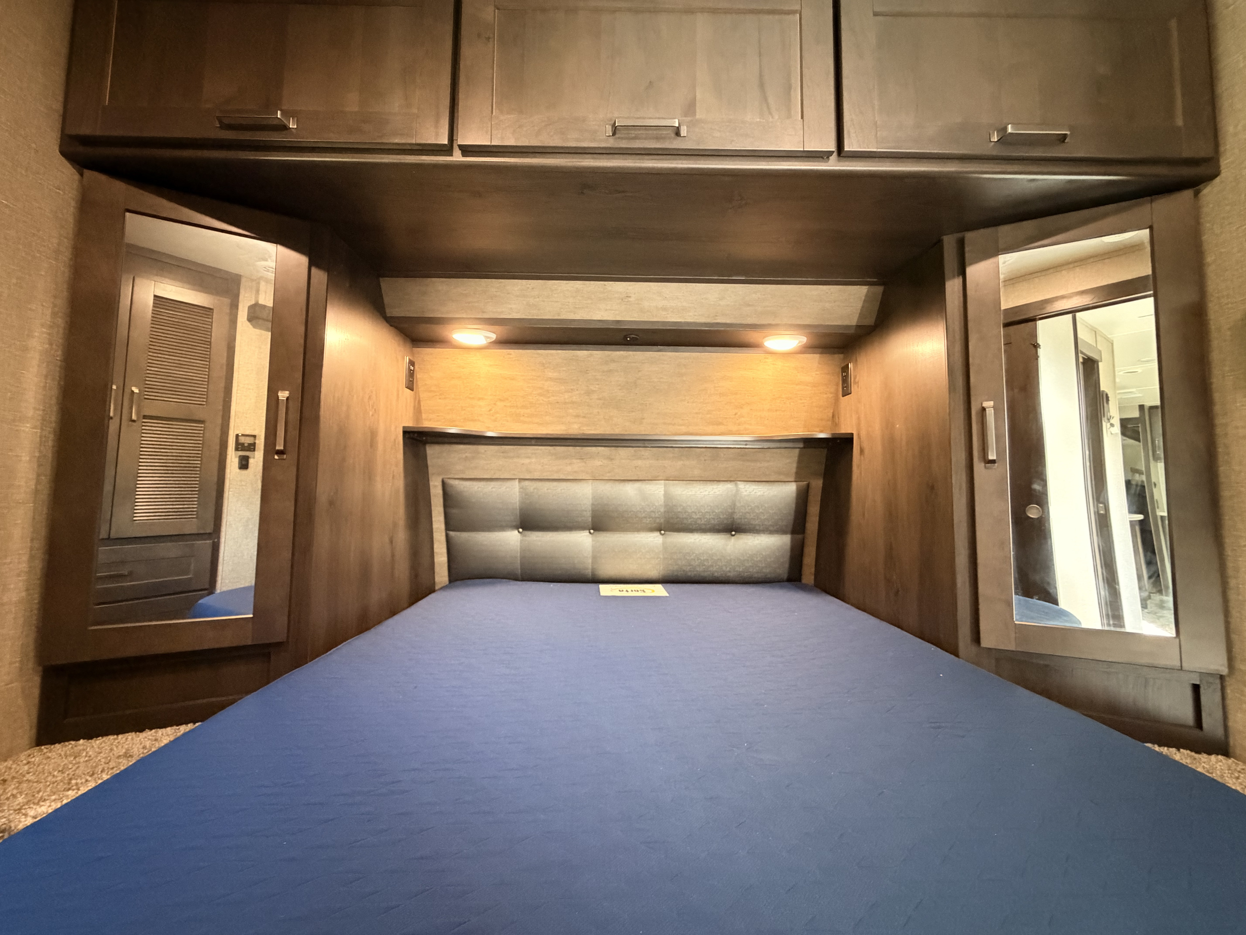 Spacious RV rental bedroom with a blue mattress, padded headboard, storage cabinets, and mirrored closet doors.. Grand Design Reflection 2022