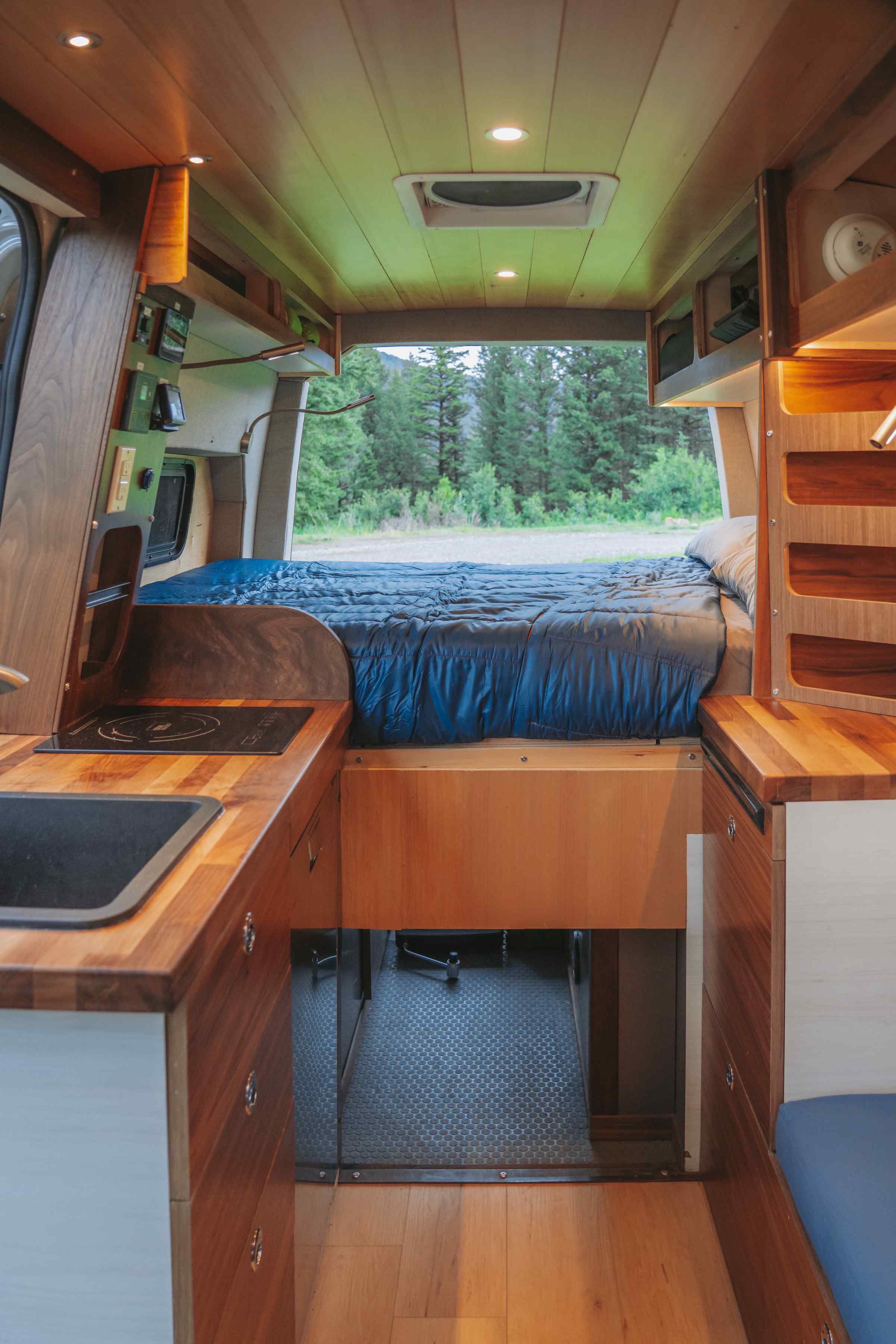 Inside an rv rental: a rustic yet modern camper van with a full bed, built-in kitchenette, wood ceiling, and forest backdrop.. Custom Mercedes Benz Sprinter 2022