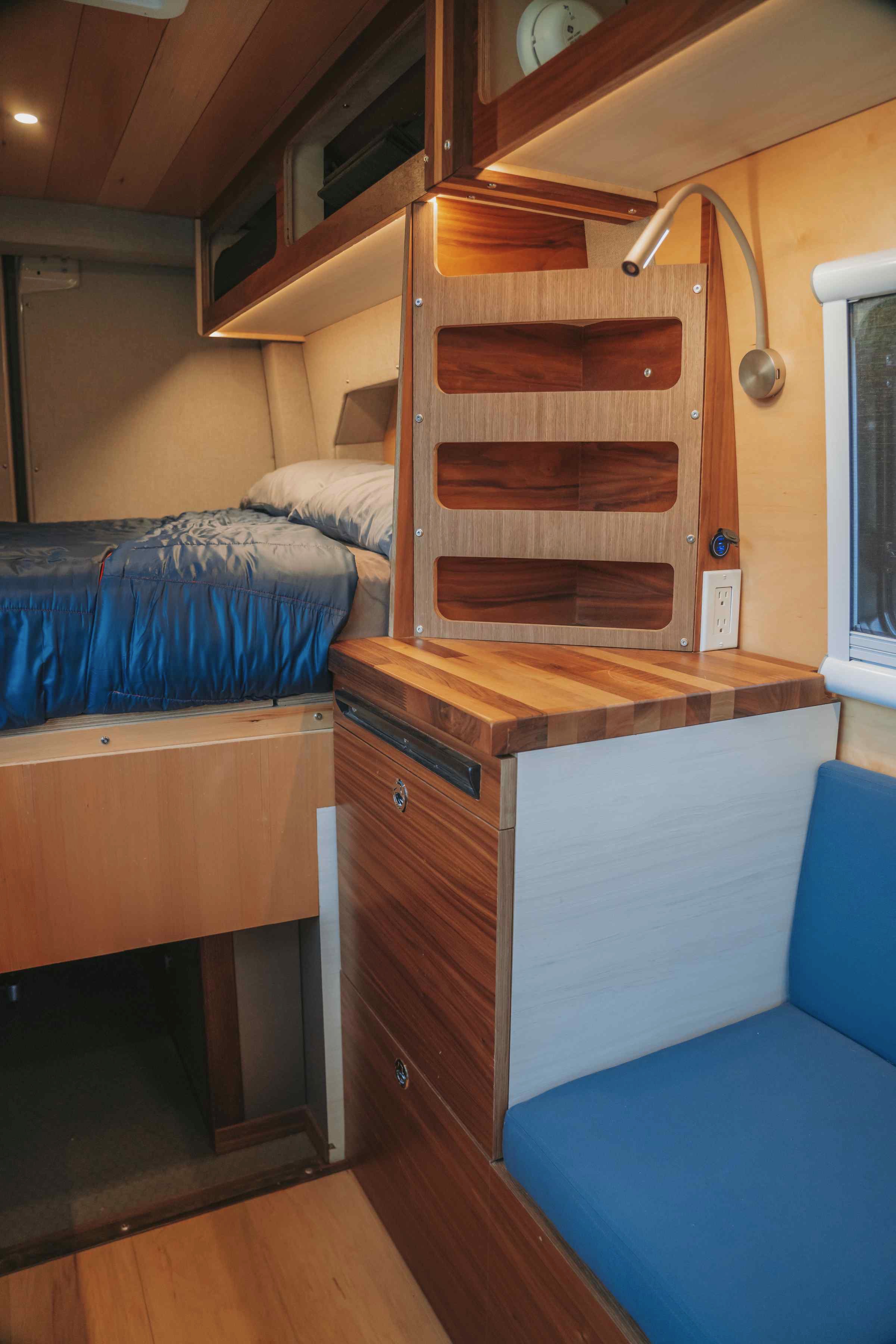 A cozy RV rental camper van interior featuring a bed, wooden storage, and blue cushioned seating.. Custom Mercedes Benz Sprinter 2022