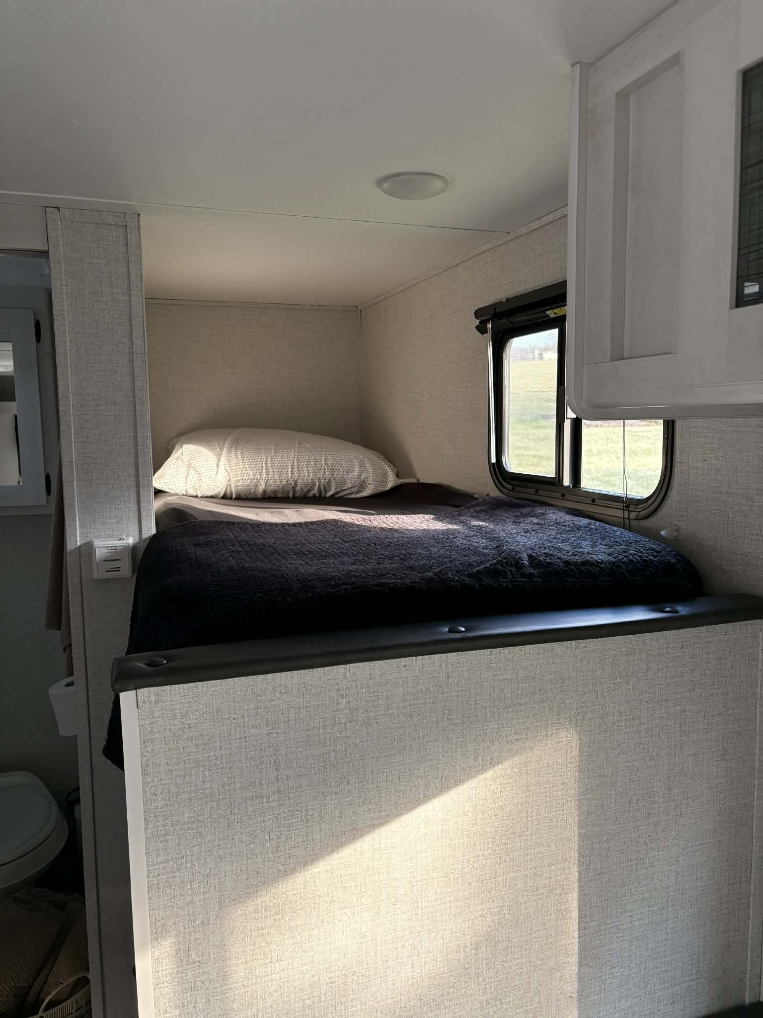 This RV rental features a compact bunk bed with dark bedding, a white pillow, and a sunny window view, perfect for camper trips.. Dutchmen Coleman Rubicon 2021