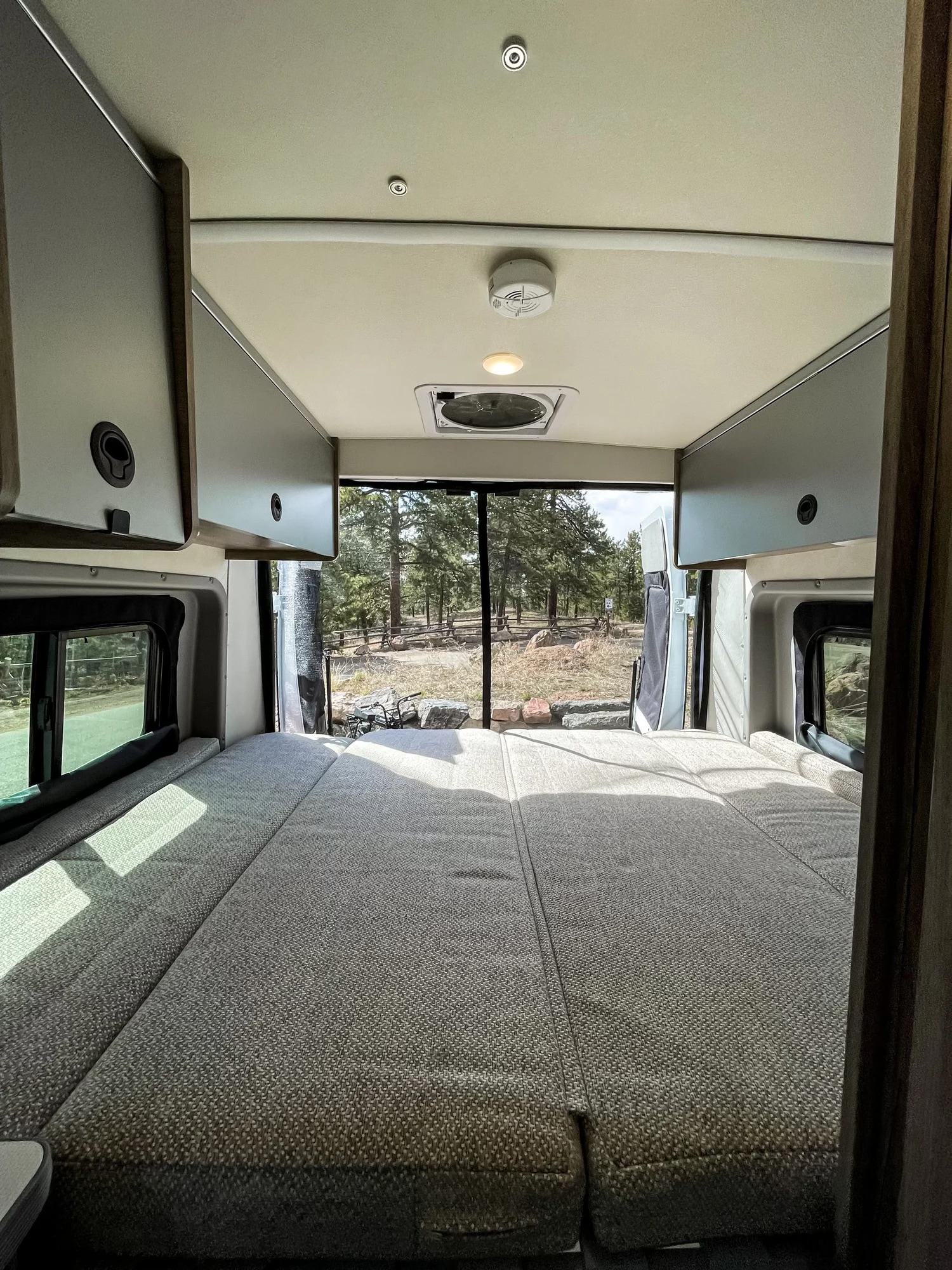 Cozy RV rental bed inside a camper van, with overhead cabinets and a beautiful nature view through the open back.. Winnebago Solis 2022