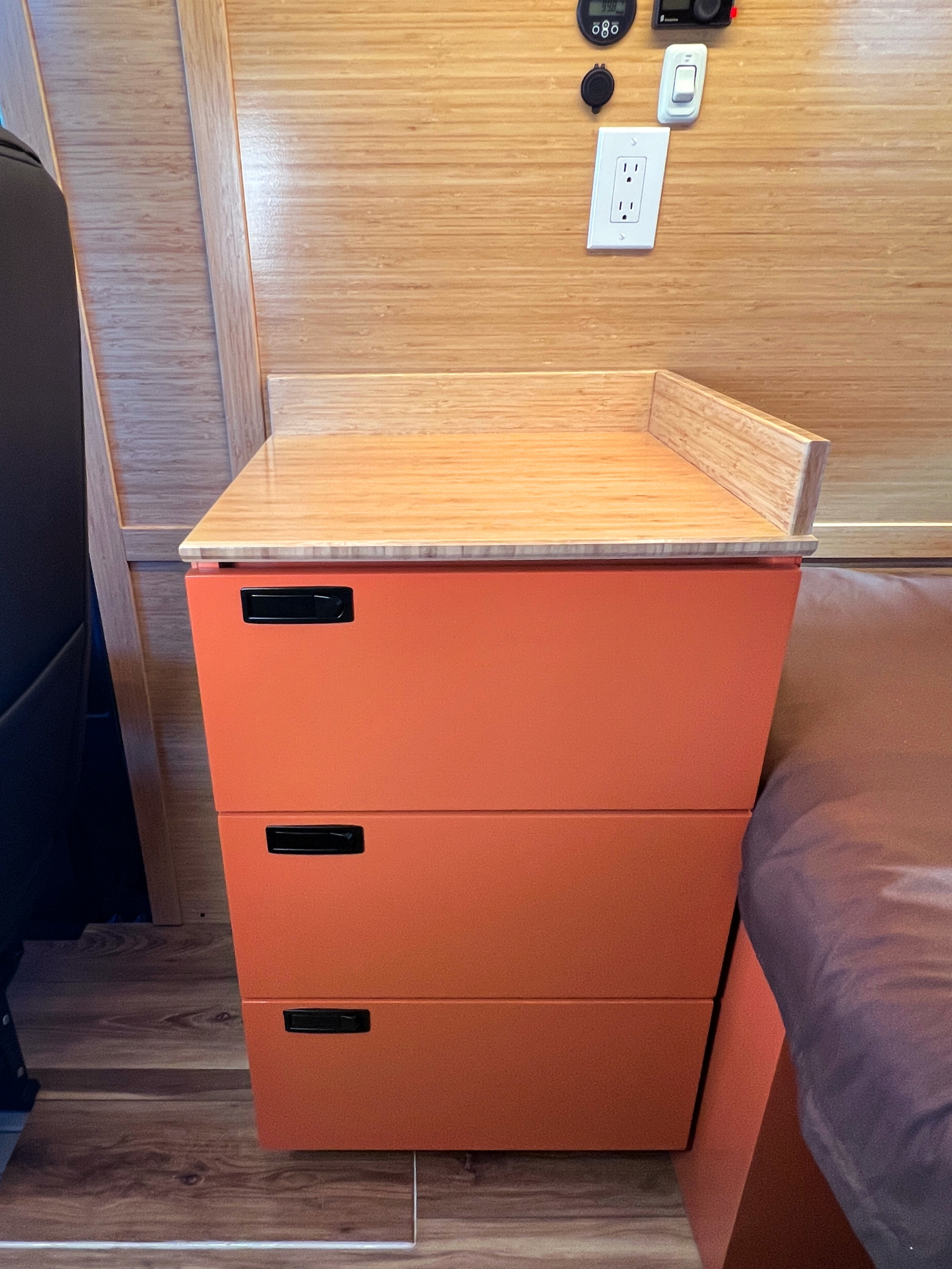 Modern orange storage cabinet with bamboo countertop, electrical outlet, and bed in a bright RV rental interior.. Mercedes-Benz Sprinter 2025