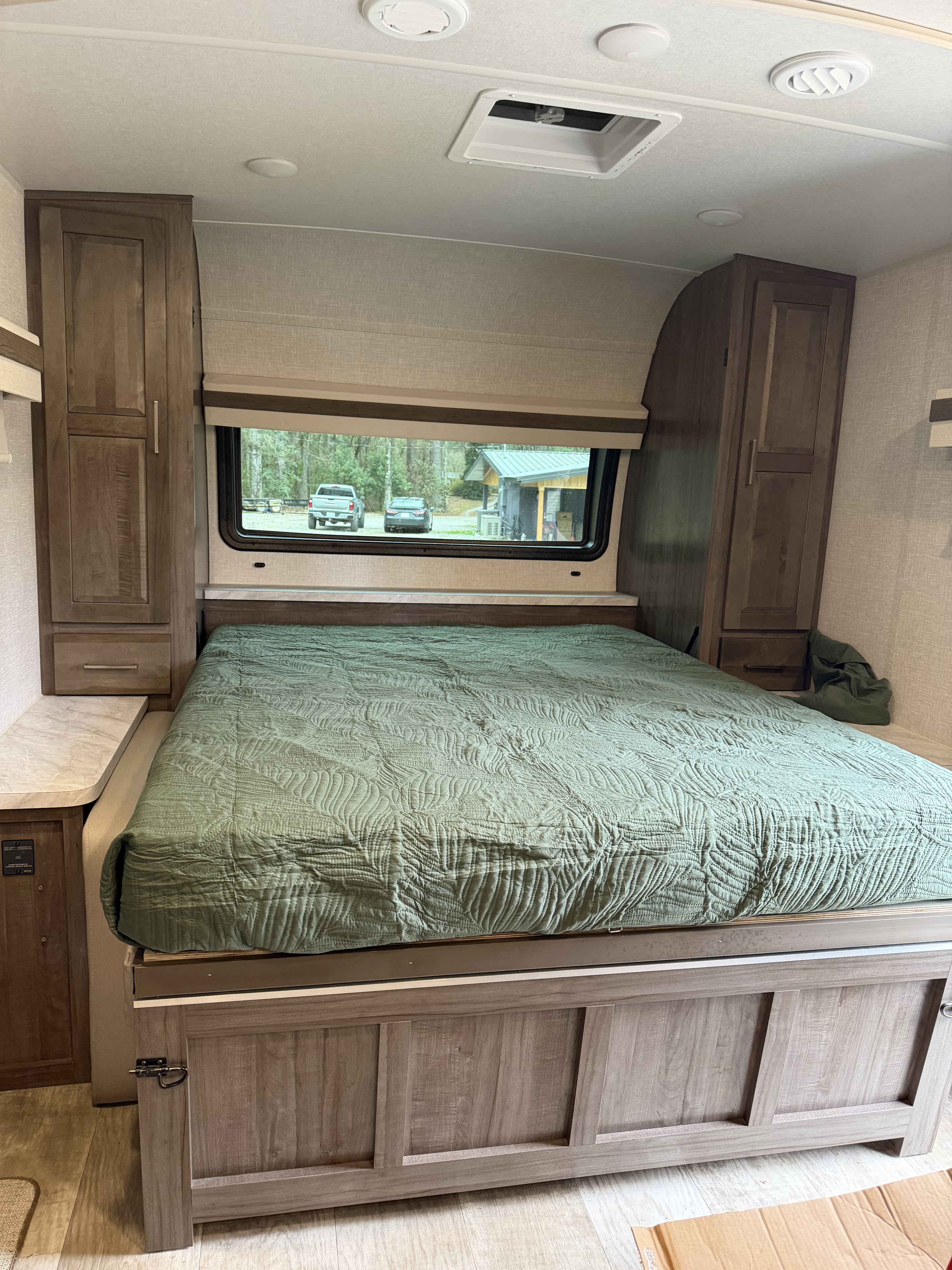 Interior of an RV rental bedroom showing a bed with a green comforter, storage cabinets, and a window view.. Forest River Rockwood Mini Lite 2023