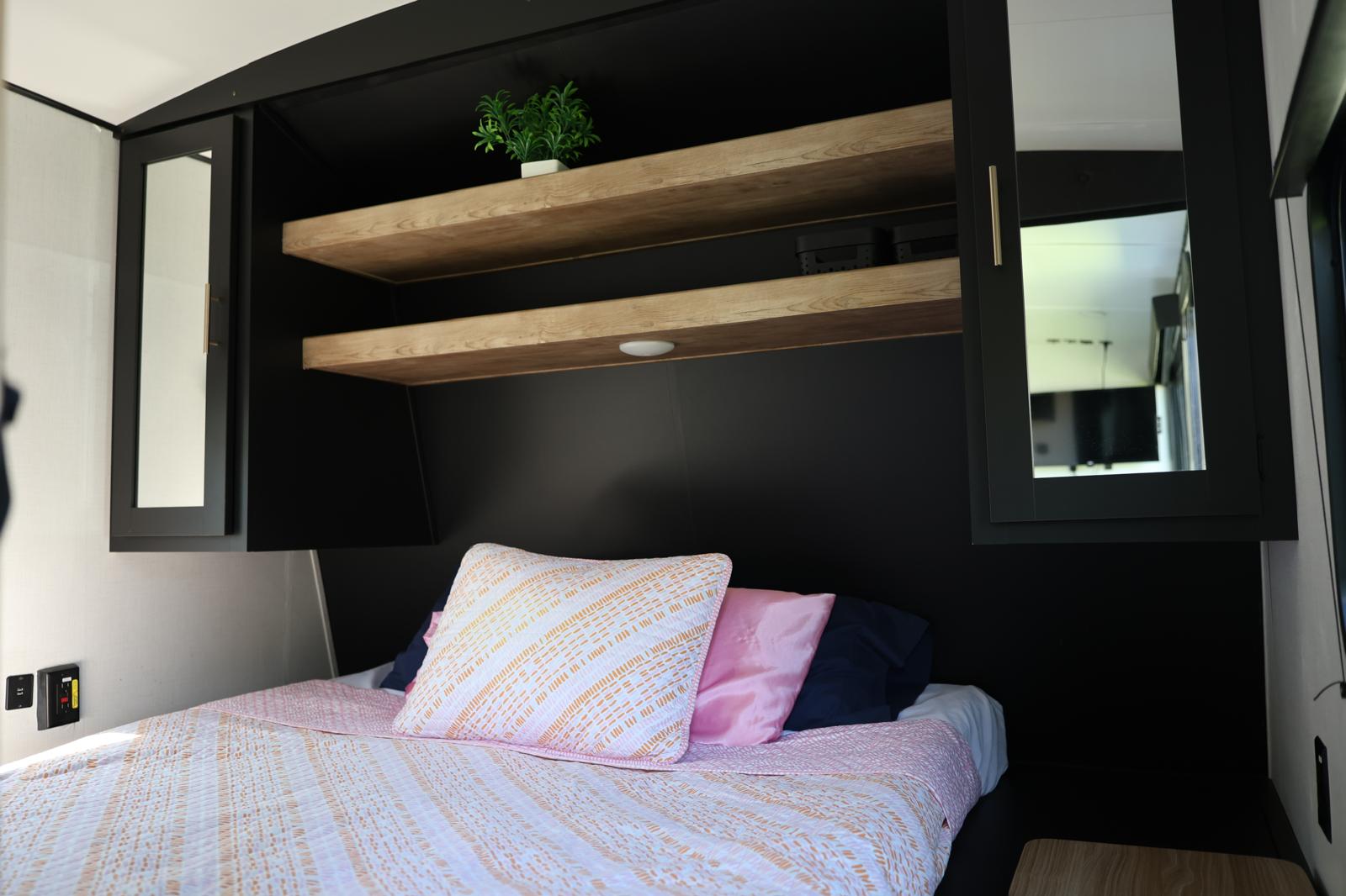 This RV rental bedroom features a comfy bed with pink bedding, wooden shelves, and mirrored cabinets above for storage.. Keystone RV Springdale 2023