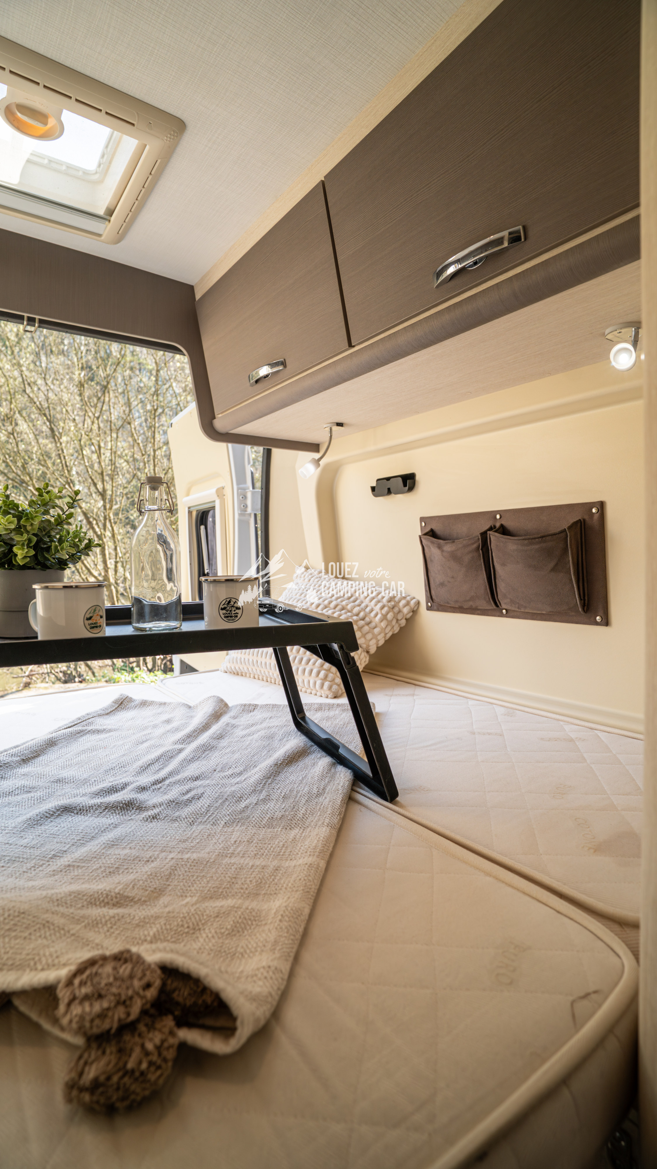 Cozy RV rental interior showing a bed with a tray table, plant, and mugs, ready for a comfortable road trip adventure.. CHALLENGER V114 2018