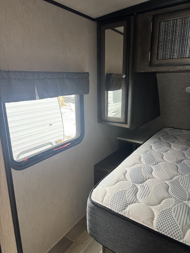 An RV rental bedroom with a comfortable mattress, window with blinds, and a mirrored cabinet, showing an emergency exit.. Heartland RVs Mallard 2019