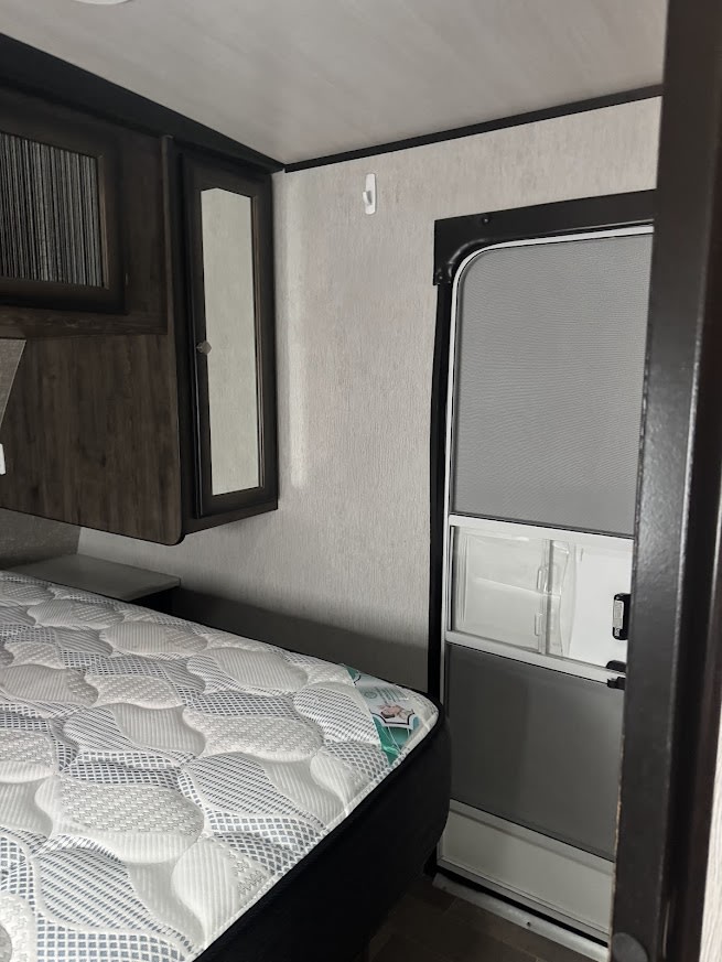 Inside an RV rental, a compact bedroom features a patterned mattress, overhead cabinet, wall mirror, and an RV screen door.. Heartland RVs Mallard 2019