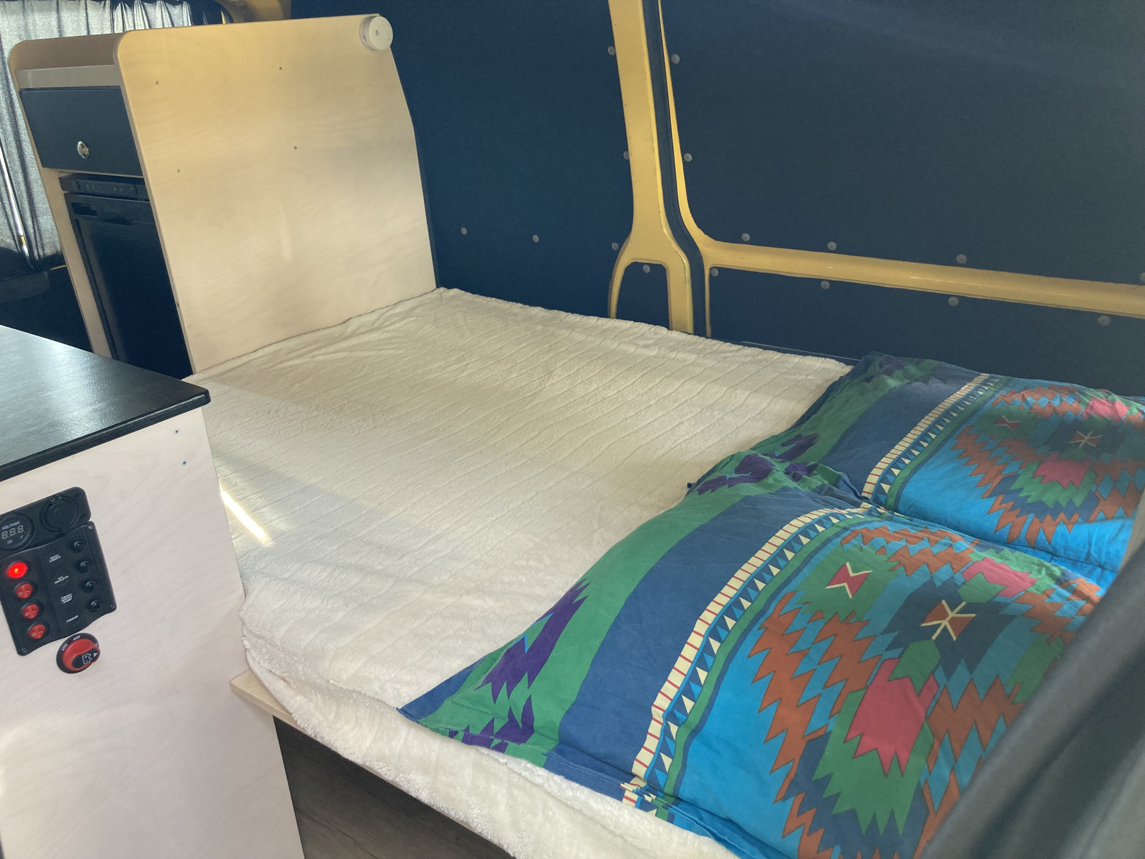 View inside an RV rental showing a bed with colorful bedding, built-in storage, and a power control panel.. Volkswagen Transporter 2010