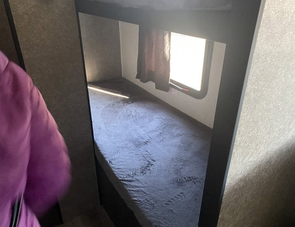 RV rental bunk bed interior, bathed in sunlight. Compact sleeping area with a window and textured blanket for travel.. Highland Ridge RV Open Range 2021