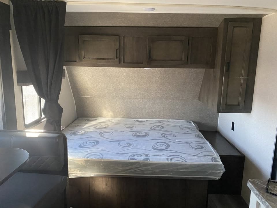 RV rental bedroom featuring a bed with a patterned mattress, overhead storage, window, and adjacent dining area.. Highland Ridge RV Open Range 2021
