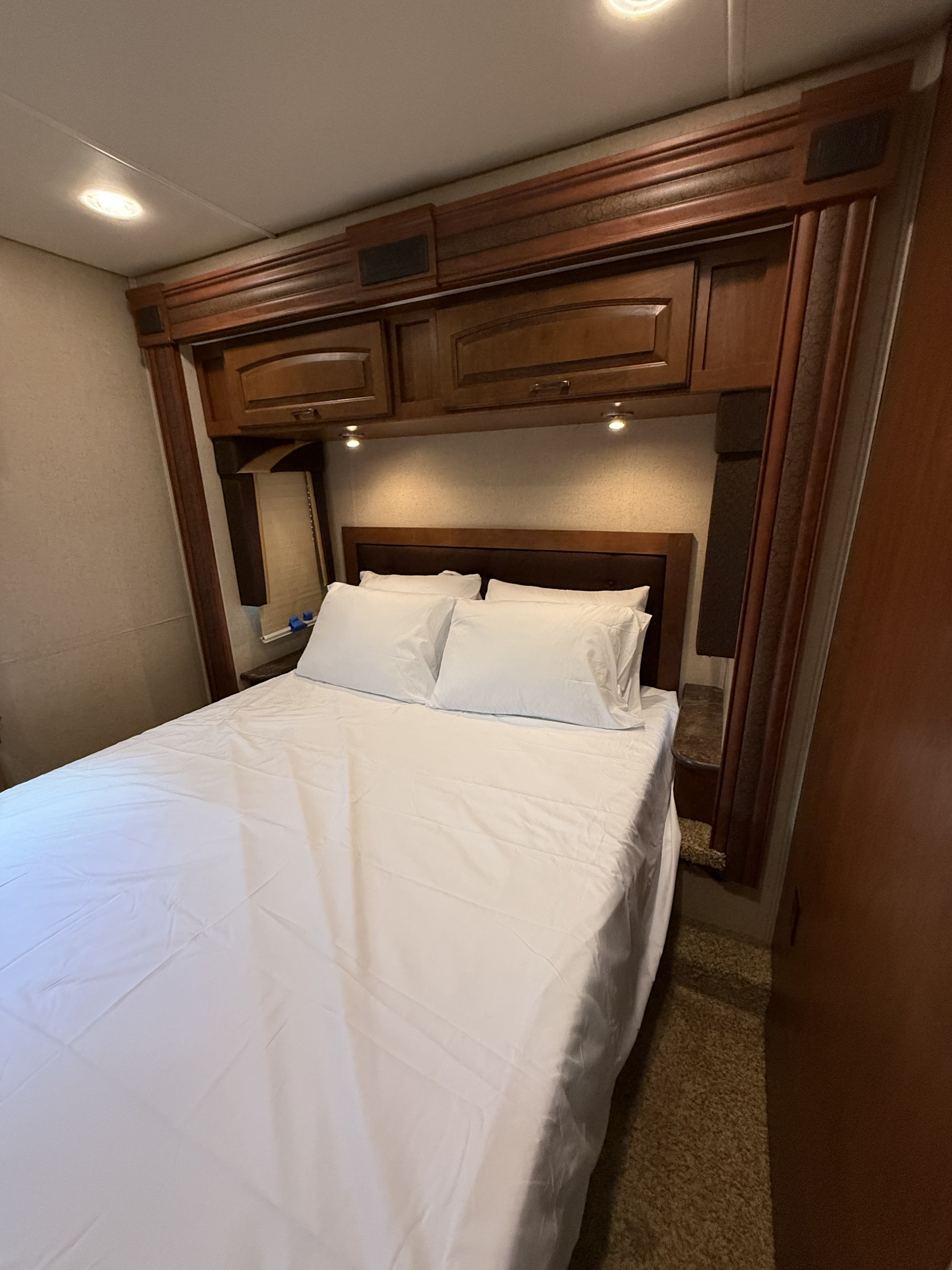 This RV rental bedroom features a comfortable bed, overhead wooden storage, and bedside lighting, perfect for a cozy trip.. Jayco Greyhawk 2015