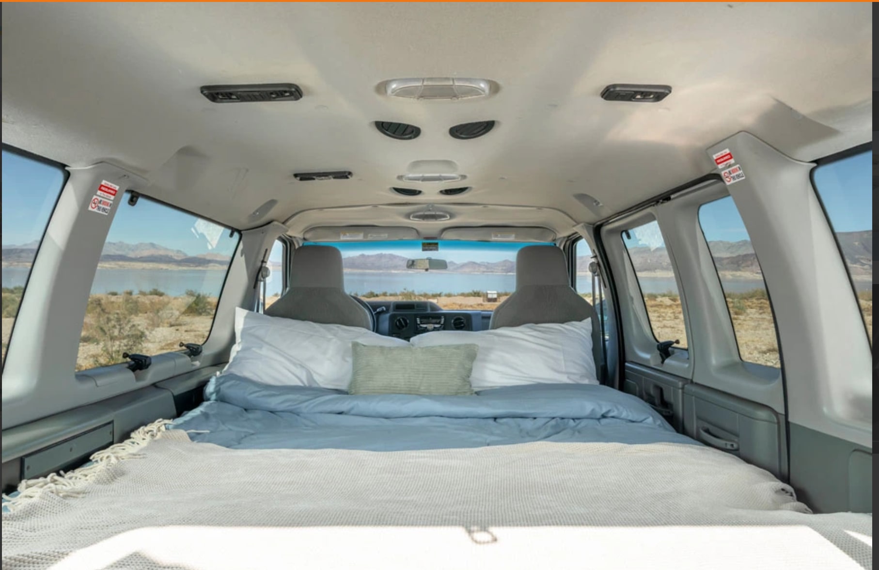 This cozy rv rental's interior features a bed facing scenic views of a lake and mountains, ideal for travel.. Ford Econoline 150 2008