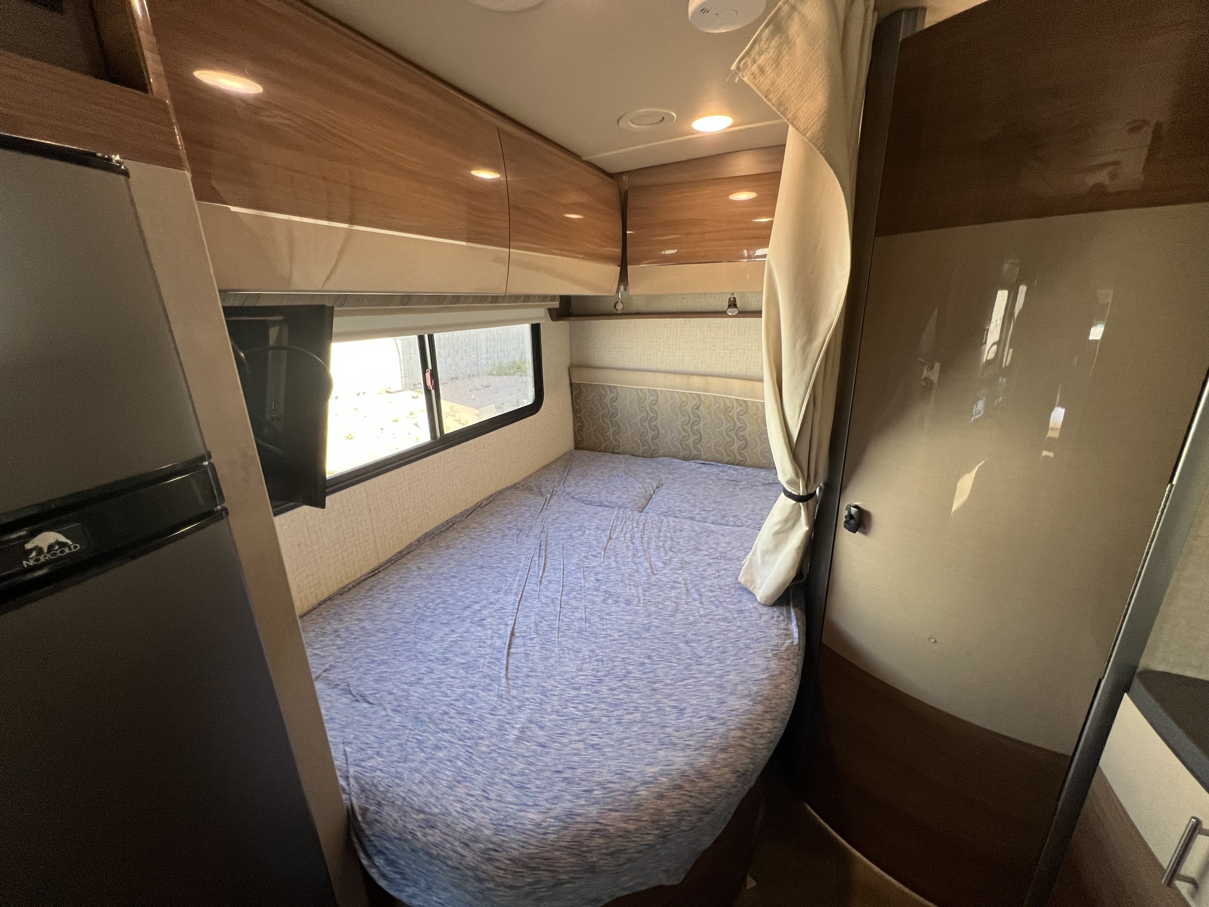 Interior of an RV rental bedroom featuring a bed with blue sheets, window, TV, Norcold fridge, and wood grain cabinets.. Winnebago View 2016