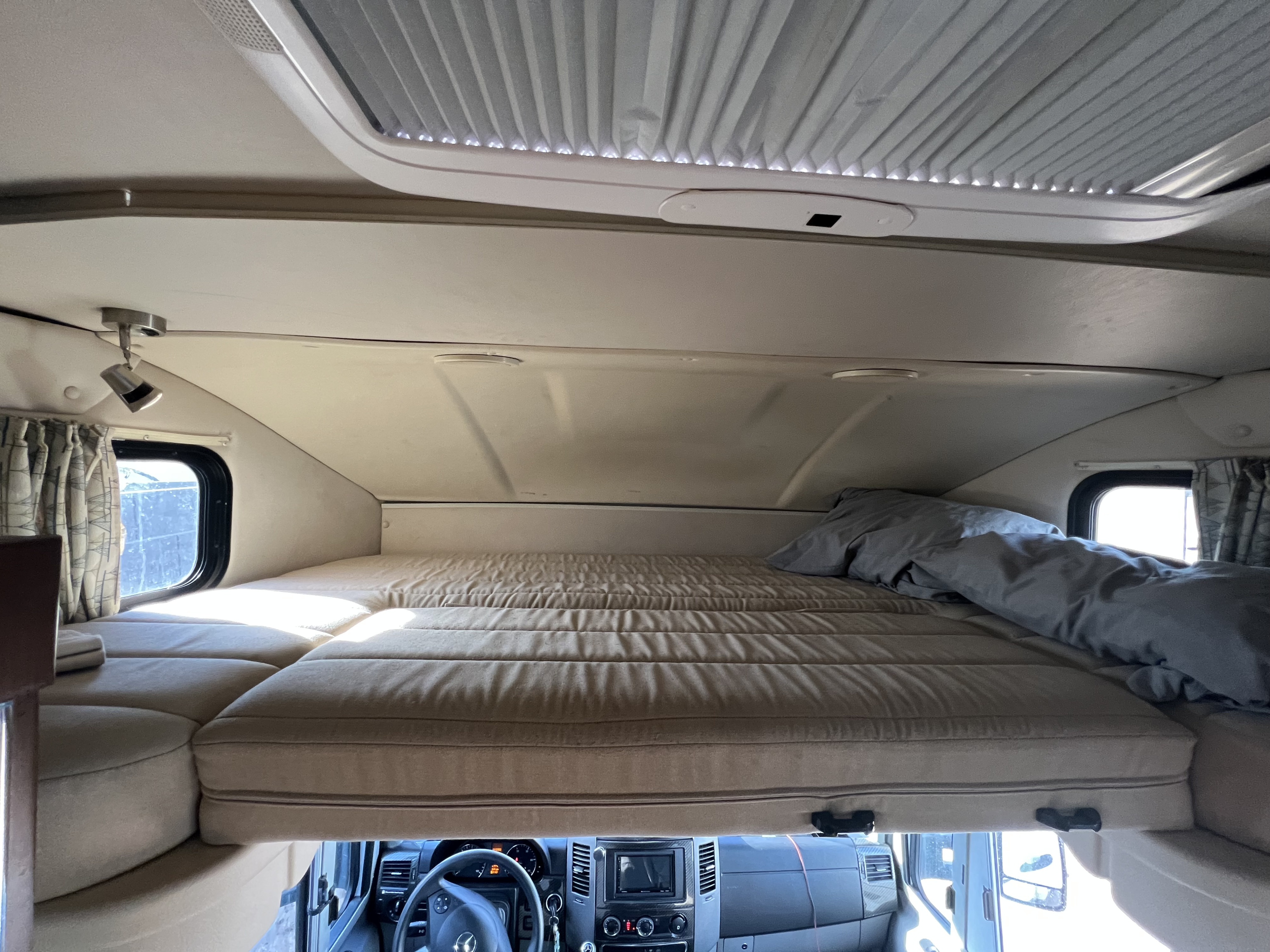 Overhead bunk bed in an RV rental, showing the comfy sleeping area above the driver's cabin with a window and ceiling vent.. Winnebago View 2016