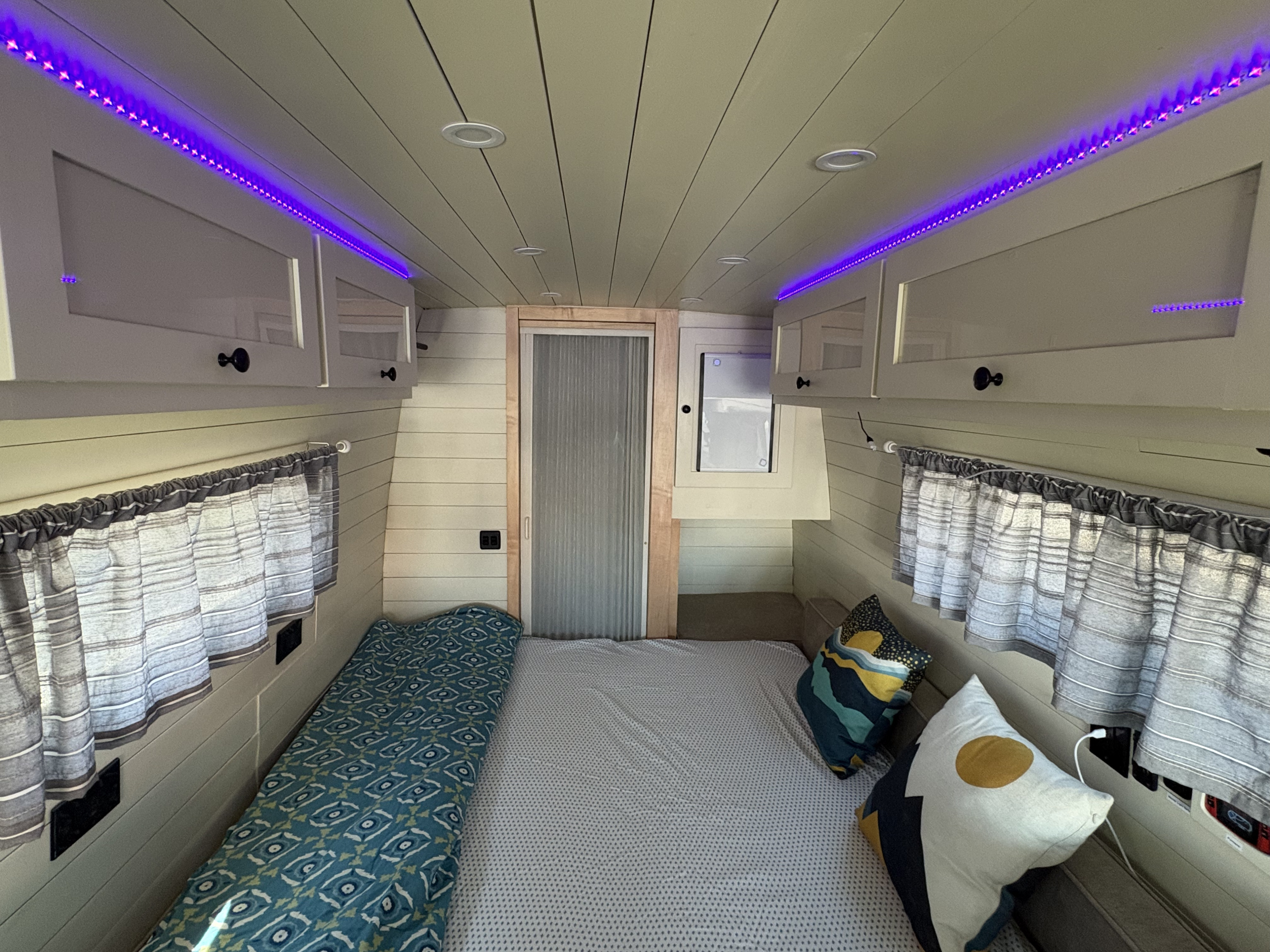 Cozy RV rental interior with a comfortable bed, vibrant purple LED lighting, overhead cabinets, and window curtains.. Mercedes-Benz Sprinter 2020