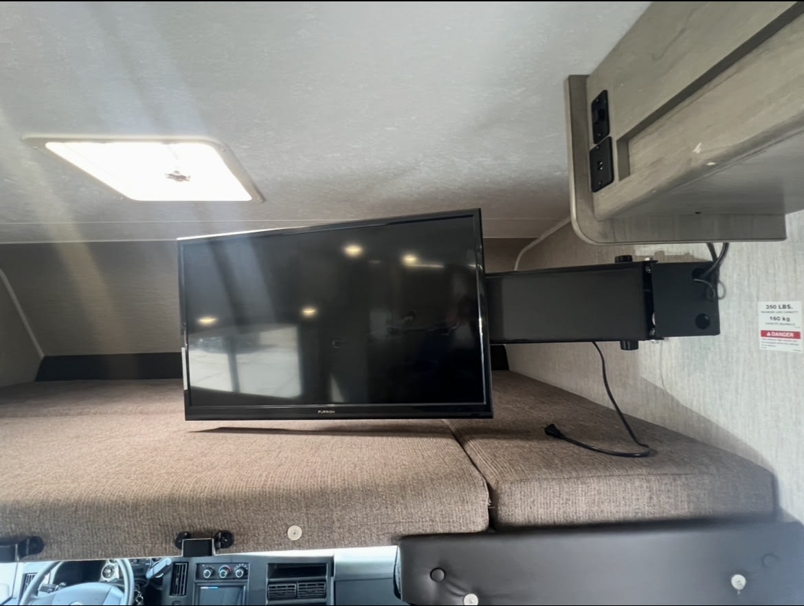 RV rental's front bunk with mounted TV on an articulating arm, overhead outlets, and ceiling light fixture.. Coachmen Freelander 2021