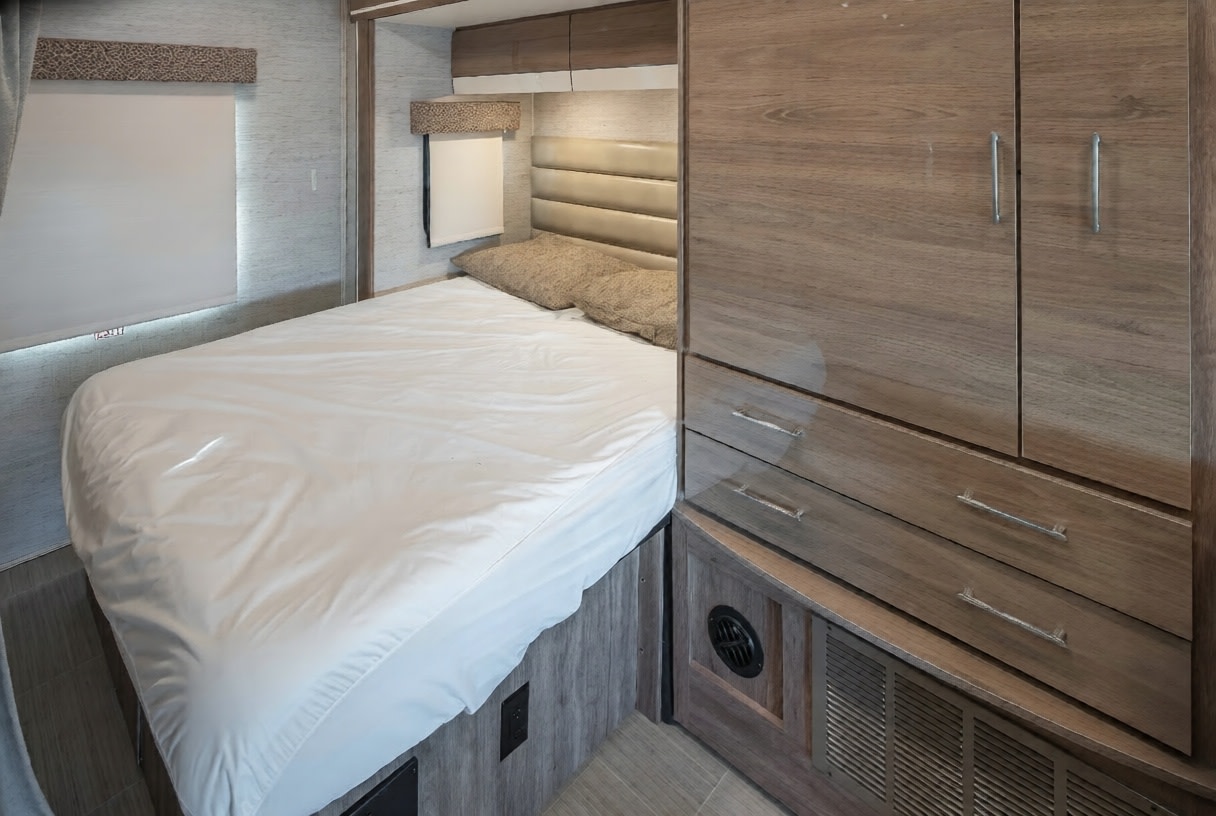 Compact RV rental bedroom featuring a white bed, upholstered headboard, window with a roller blind, and sleek wooden cabinetry.. Jayco Melbourne Prestige 2021