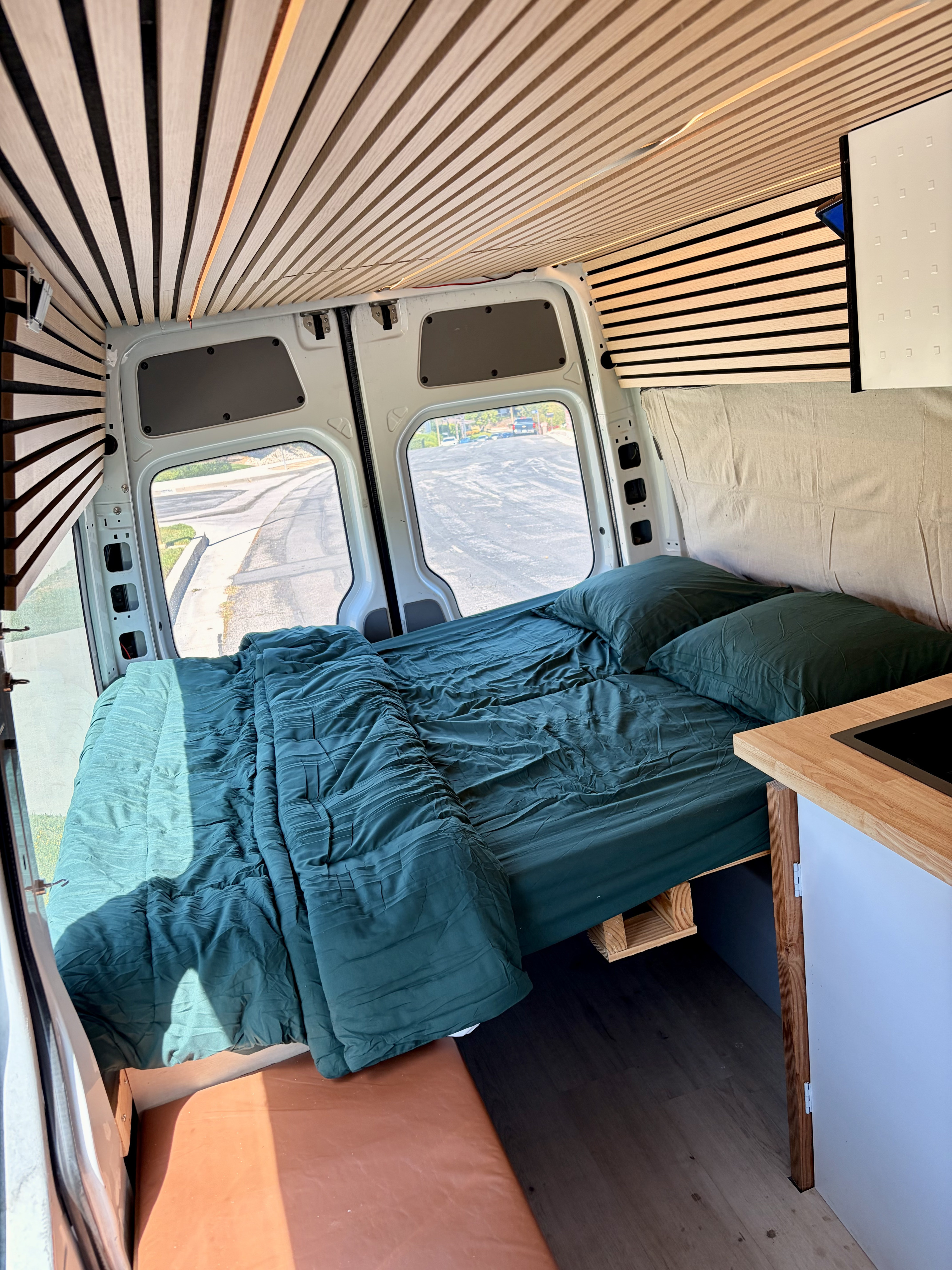 Cozy RV rental interior with a comfy dark green bed, unique wooden slat ceiling, and kitchen counter. Back doors open to a street view.. Mercedes-Benz Sprinter 2008