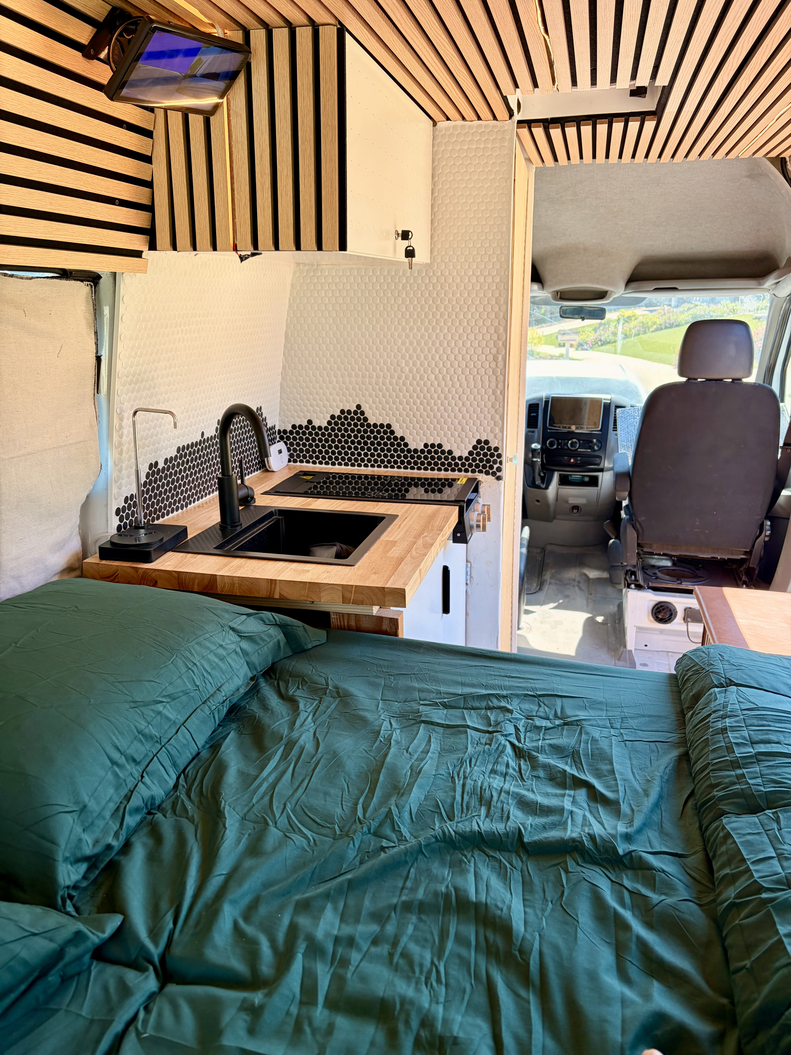 Cozy dark green bed inside a modern camper van rv rental with a kitchen, wooden accents, and driver's area visible.. Mercedes-Benz Sprinter 2008