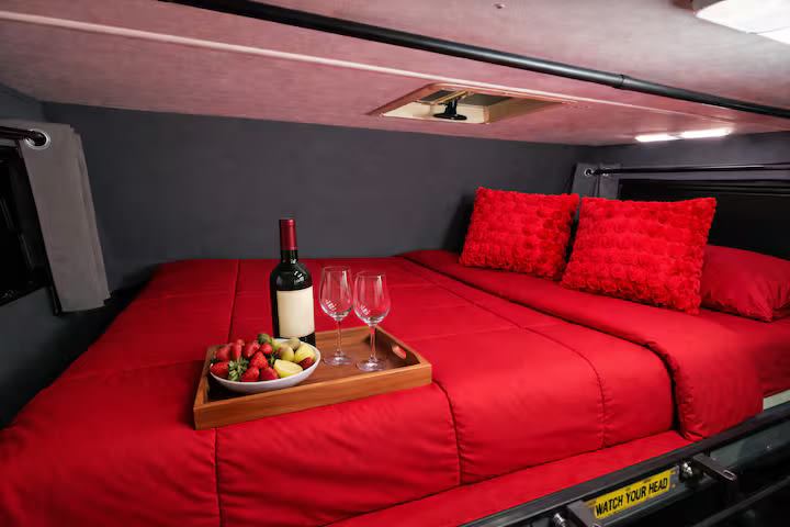 Cozy RV rental bed with red bedding, wine, and fruit on a tray. Perfect for a romantic getaway in a compact camper.. Thor Majestic 2017