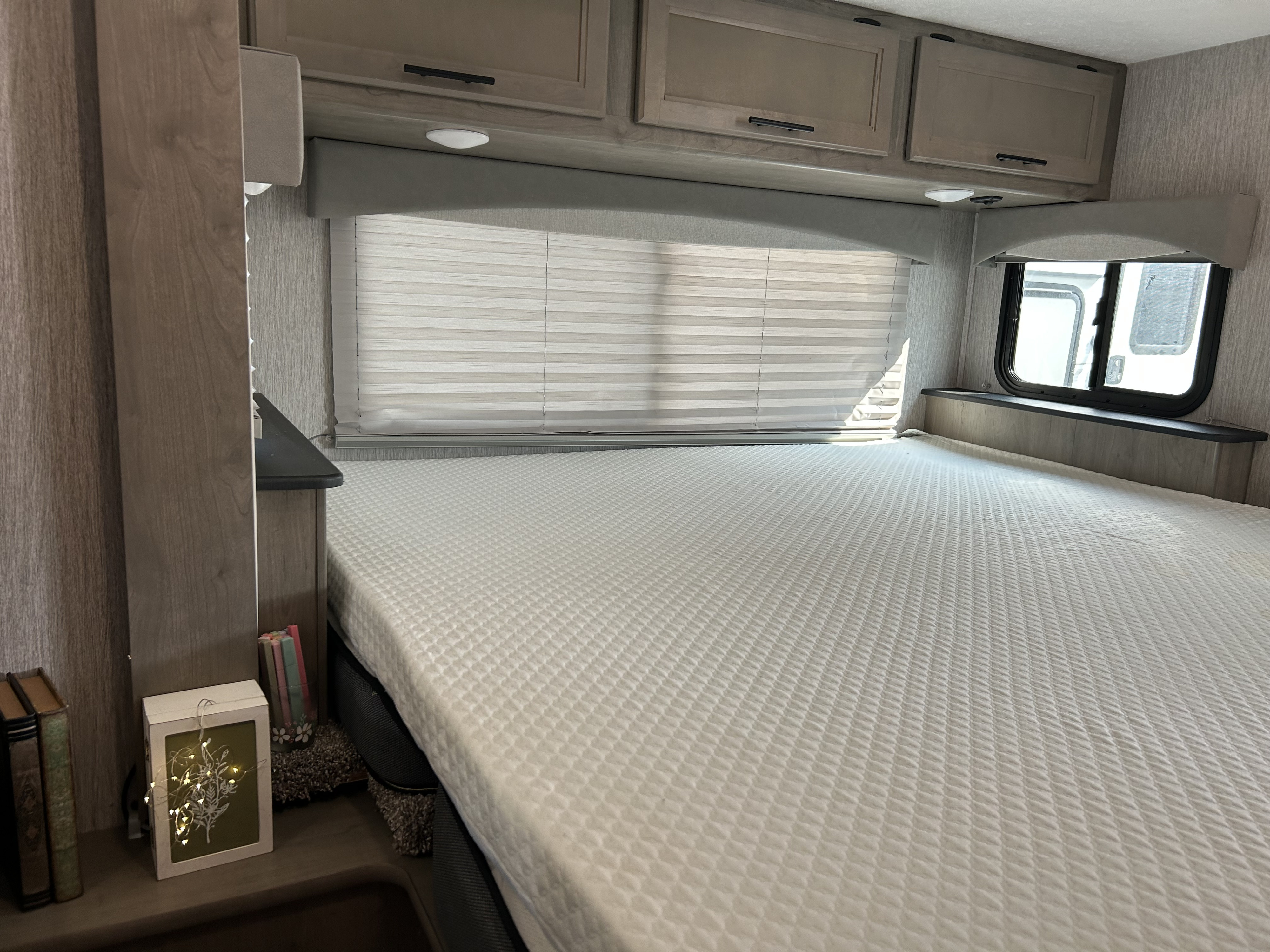 This RV rental bedroom features a comfortable, large white mattress, overhead cabinets, and windows with blinds for natural light.. Coachmen Freelander 2021