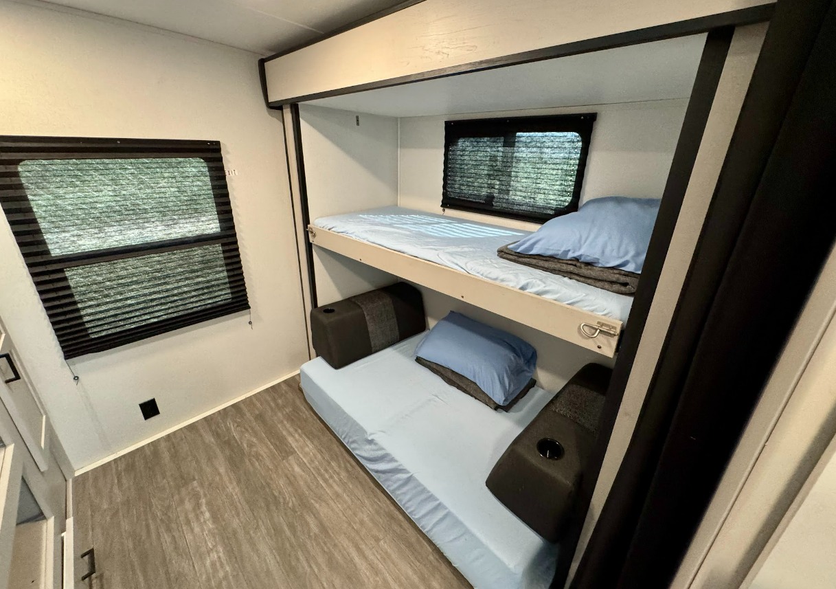 Inside an RV rental: compact bunk beds with light blue sheets & pillows, windows with blinds, and wood-look flooring.. Mallard M32 2024
