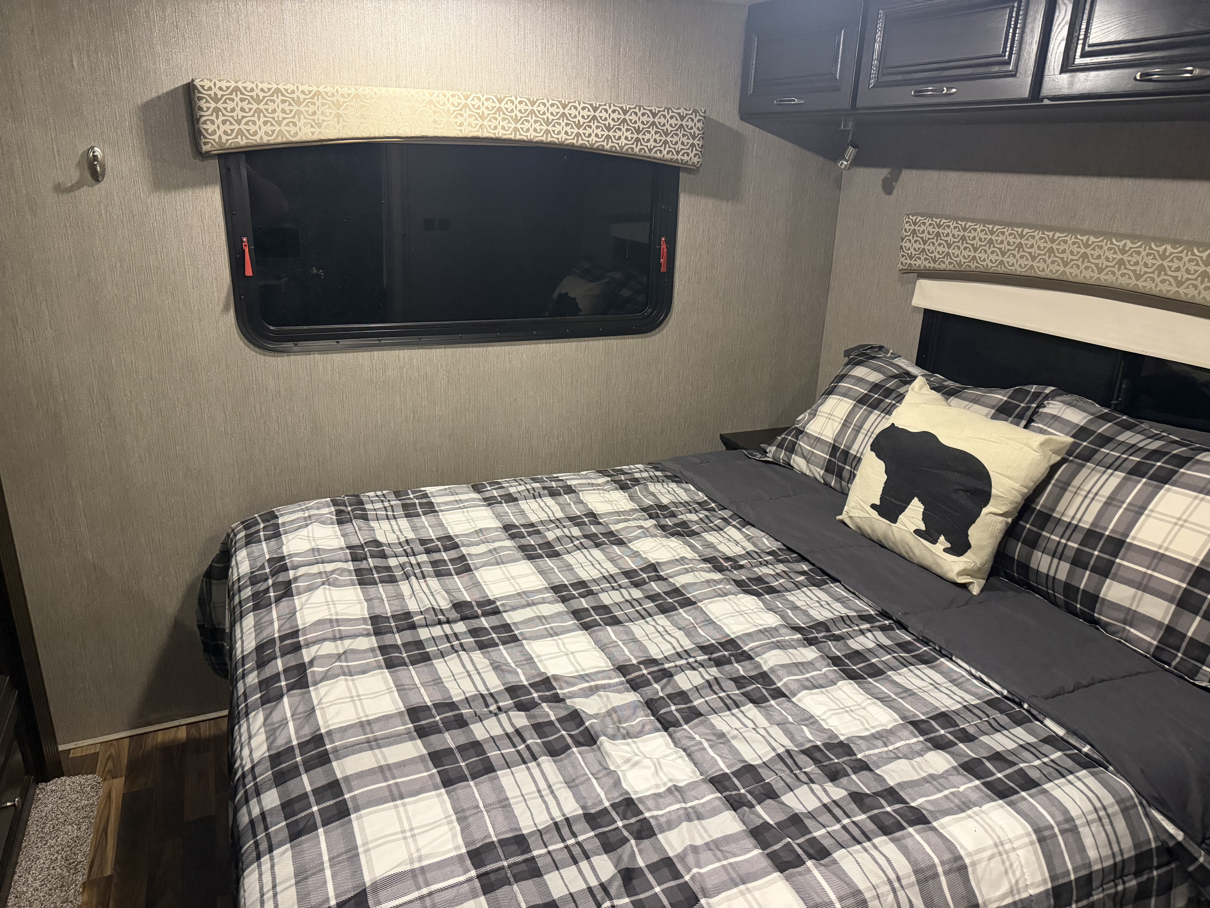 Inside a cozy RV rental bedroom: black & white plaid bed, bear pillow, dark cabinets, and textured walls.. Nexus RV Phantom 2020