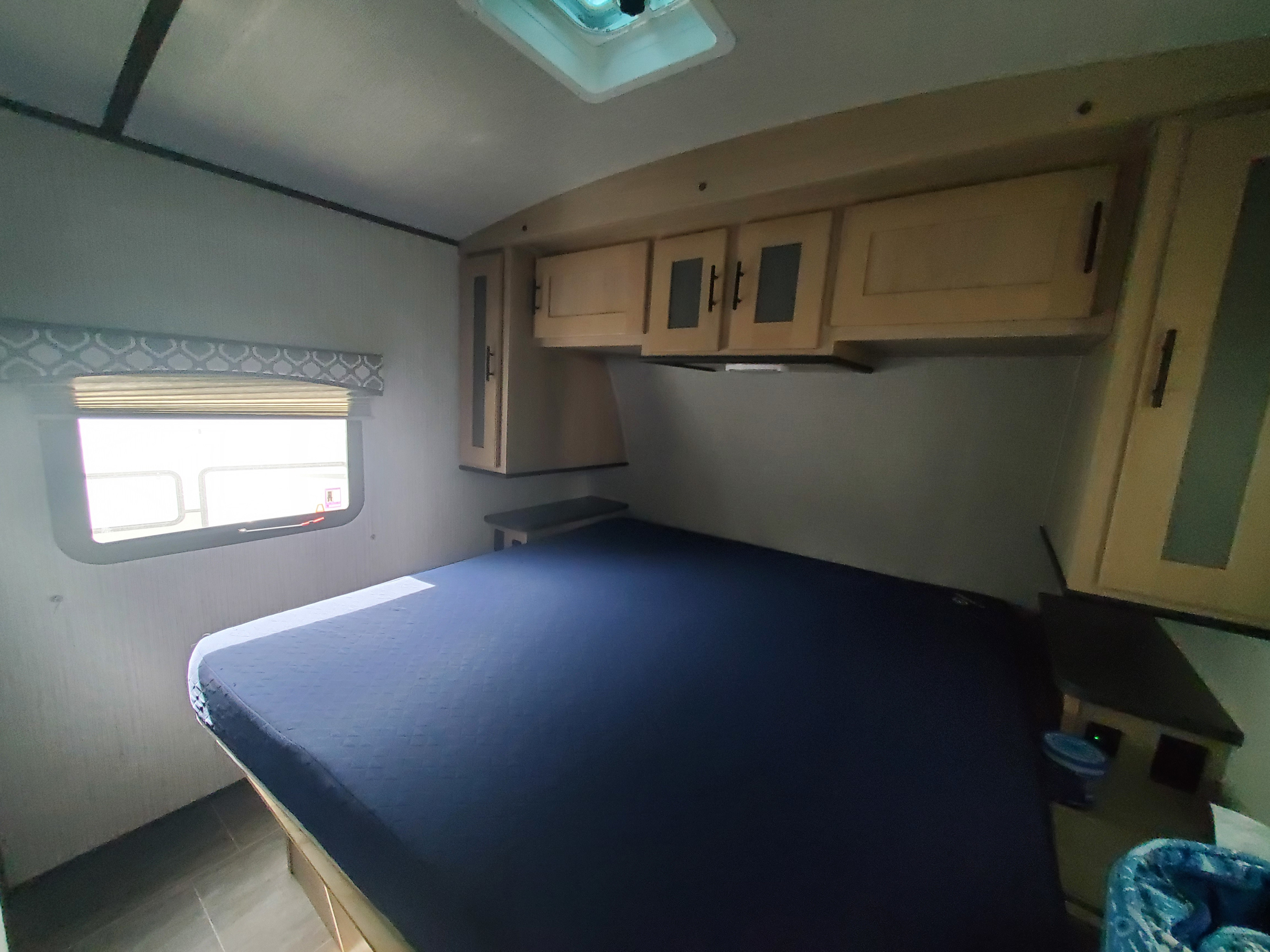 **RV rental bedroom** interior with a dark blue bed, light wood overhead cabinets, and a window with blinds.. Cruiser RV Radiance 2021